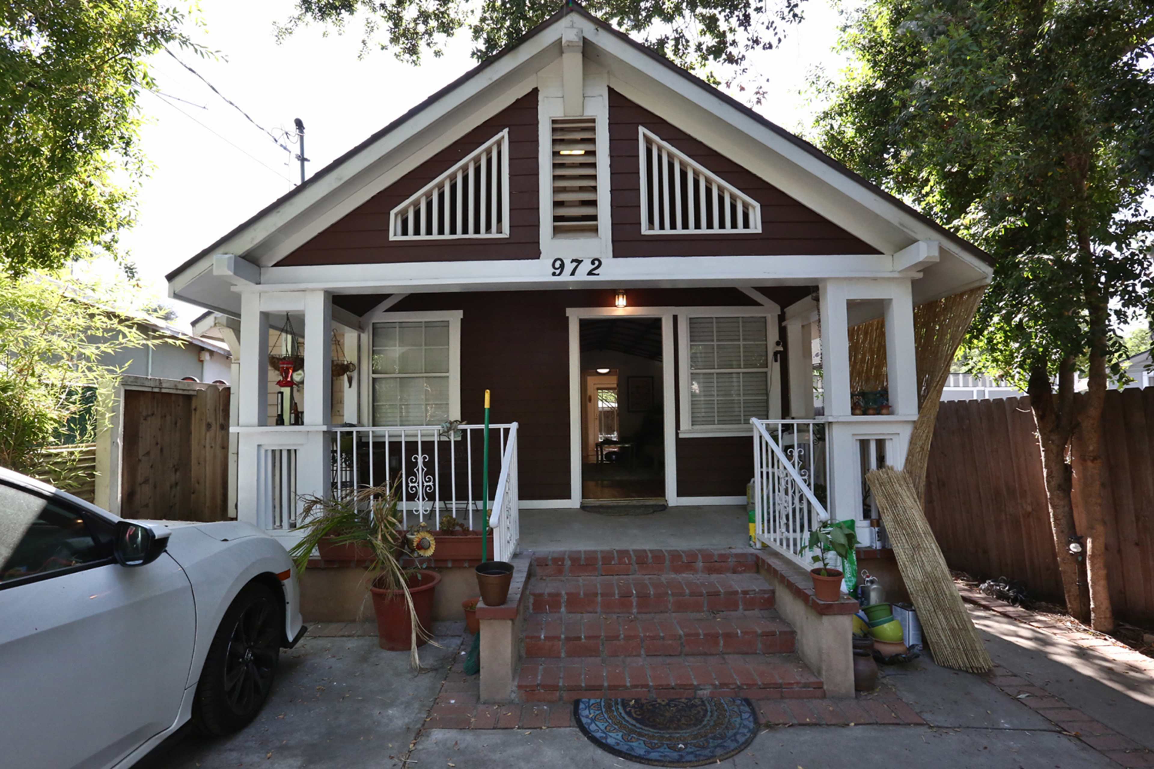The image shows a single-story brown house with a white porch and steps, featuring a front door and several plants on the porch.