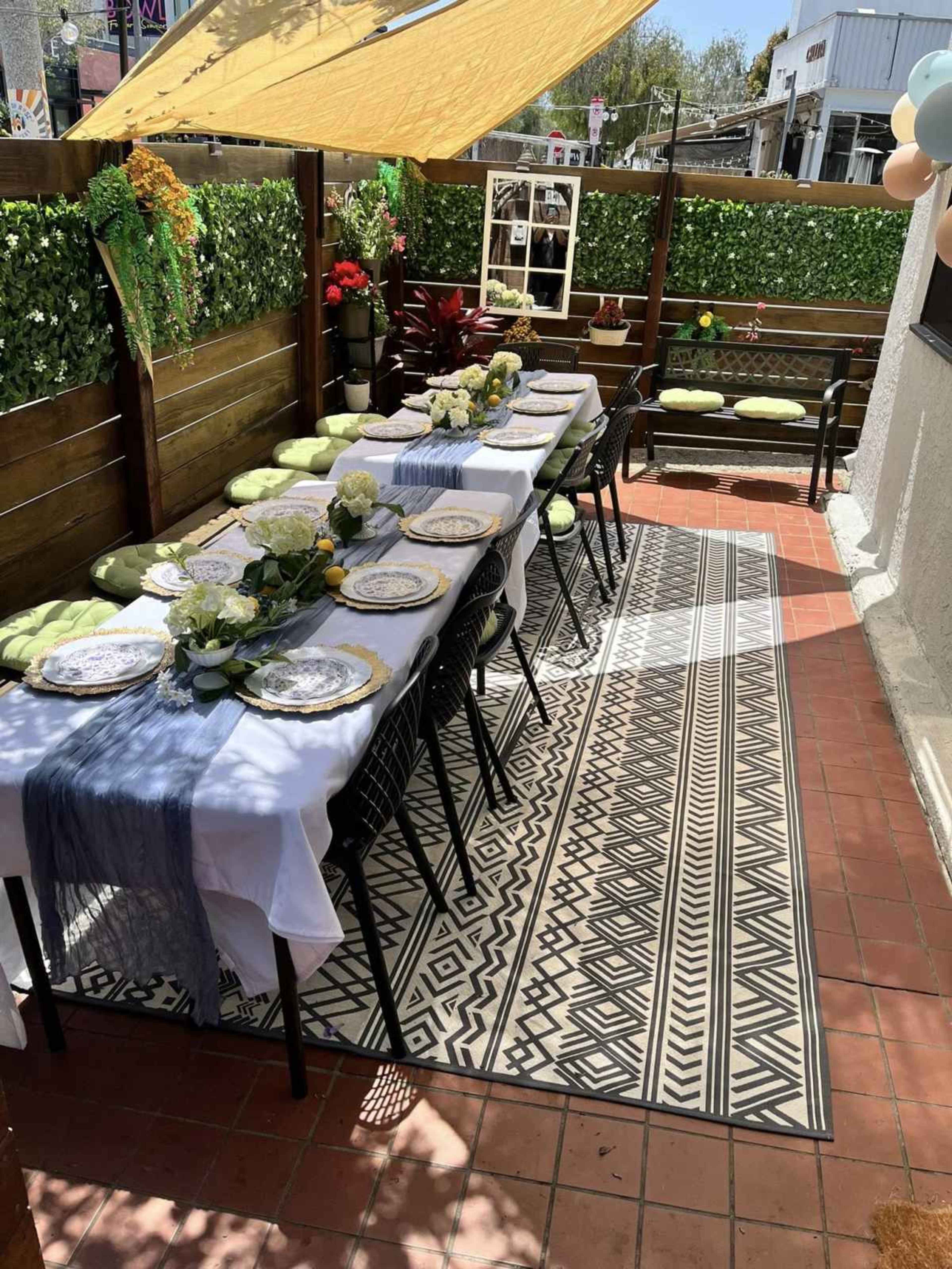 A long dining table is set with decorative tableware under an awning on a patio, surrounded by potted plants and a patterned rug.
