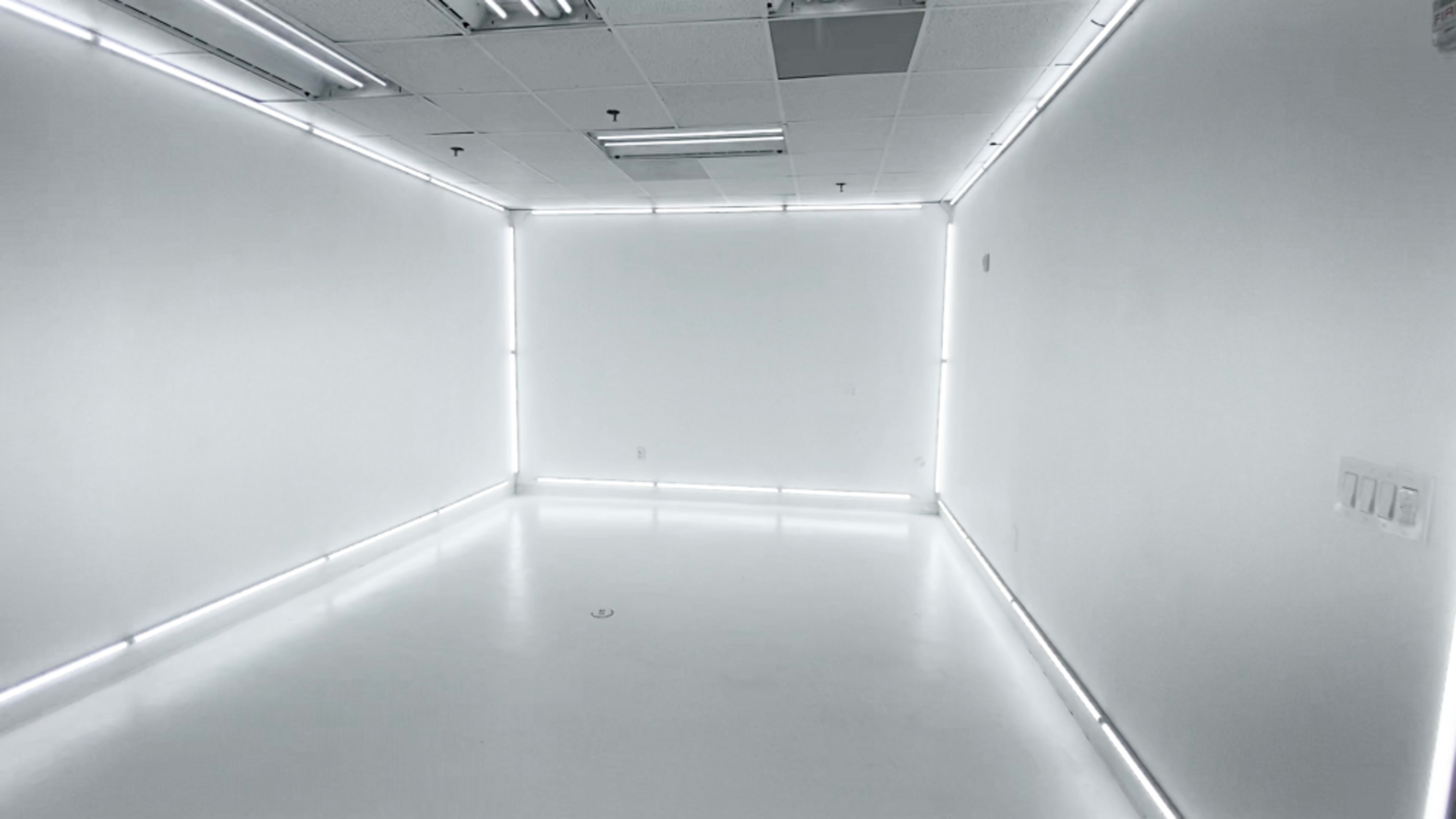 The image shows a stark, empty room with bright white walls and illuminated corners created by LED lights.
