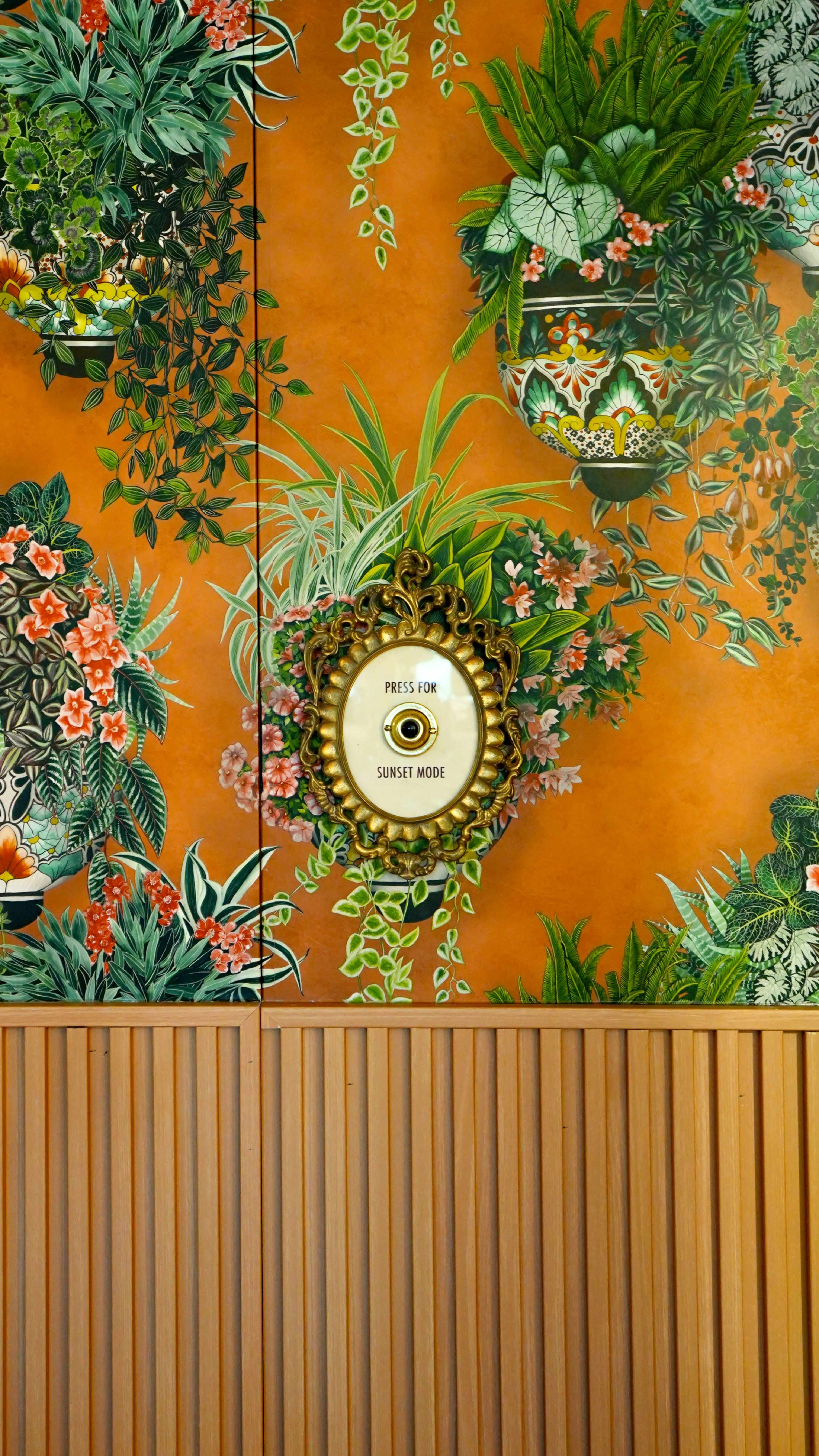The image features an orange wall adorned with vibrant plant illustrations, with a decorative button labeled "PRESS FOR SUNSET MODE" centrally positioned.