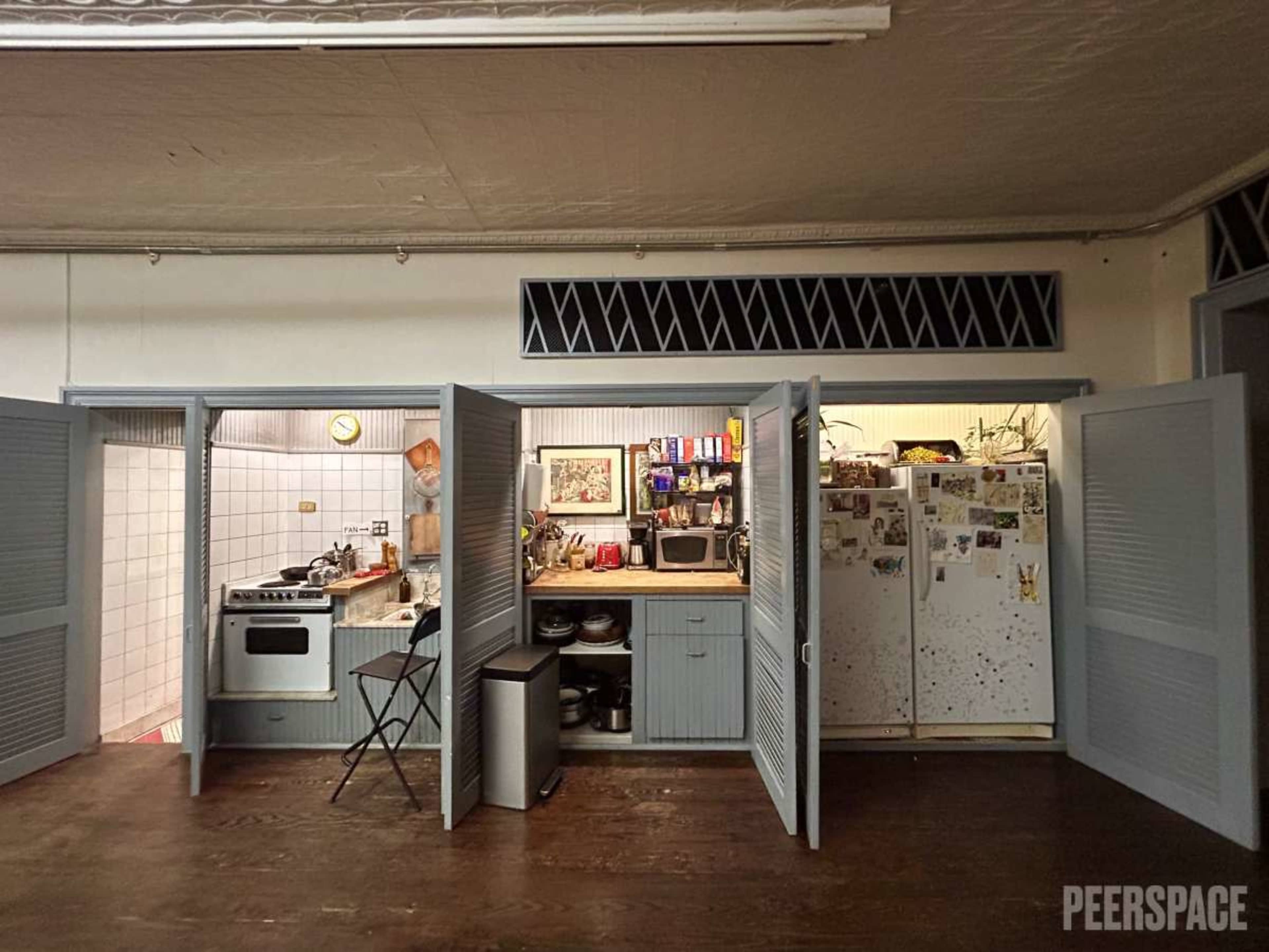 The image shows a compact kitchen with blue shutters, featuring a stove, microwave, refrigerator, and various kitchen items organized on shelves.