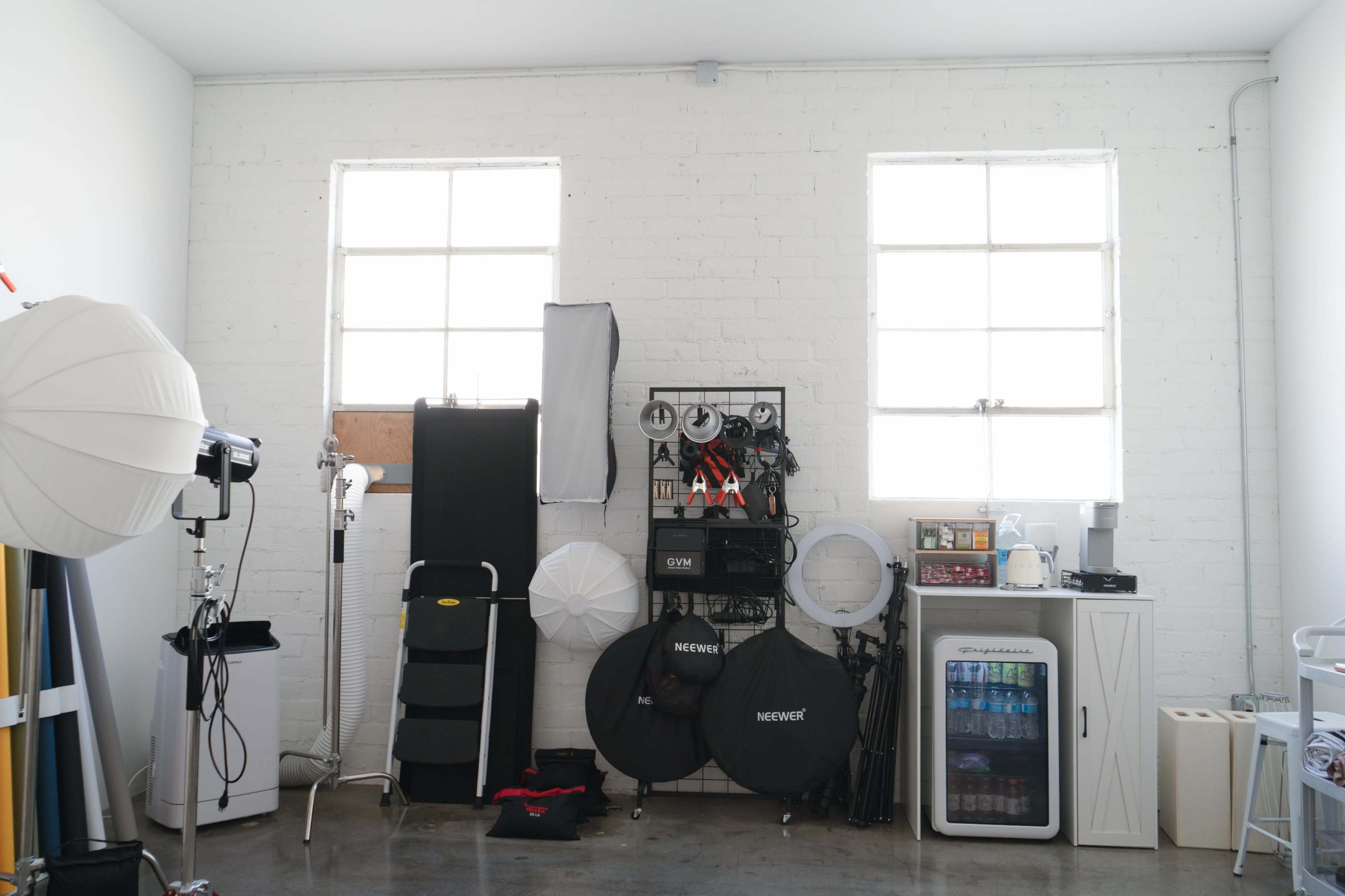 The image shows a bright studio space with large windows, featuring photography equipment, softboxes, a mini fridge, and various backdrops arranged along the wall.