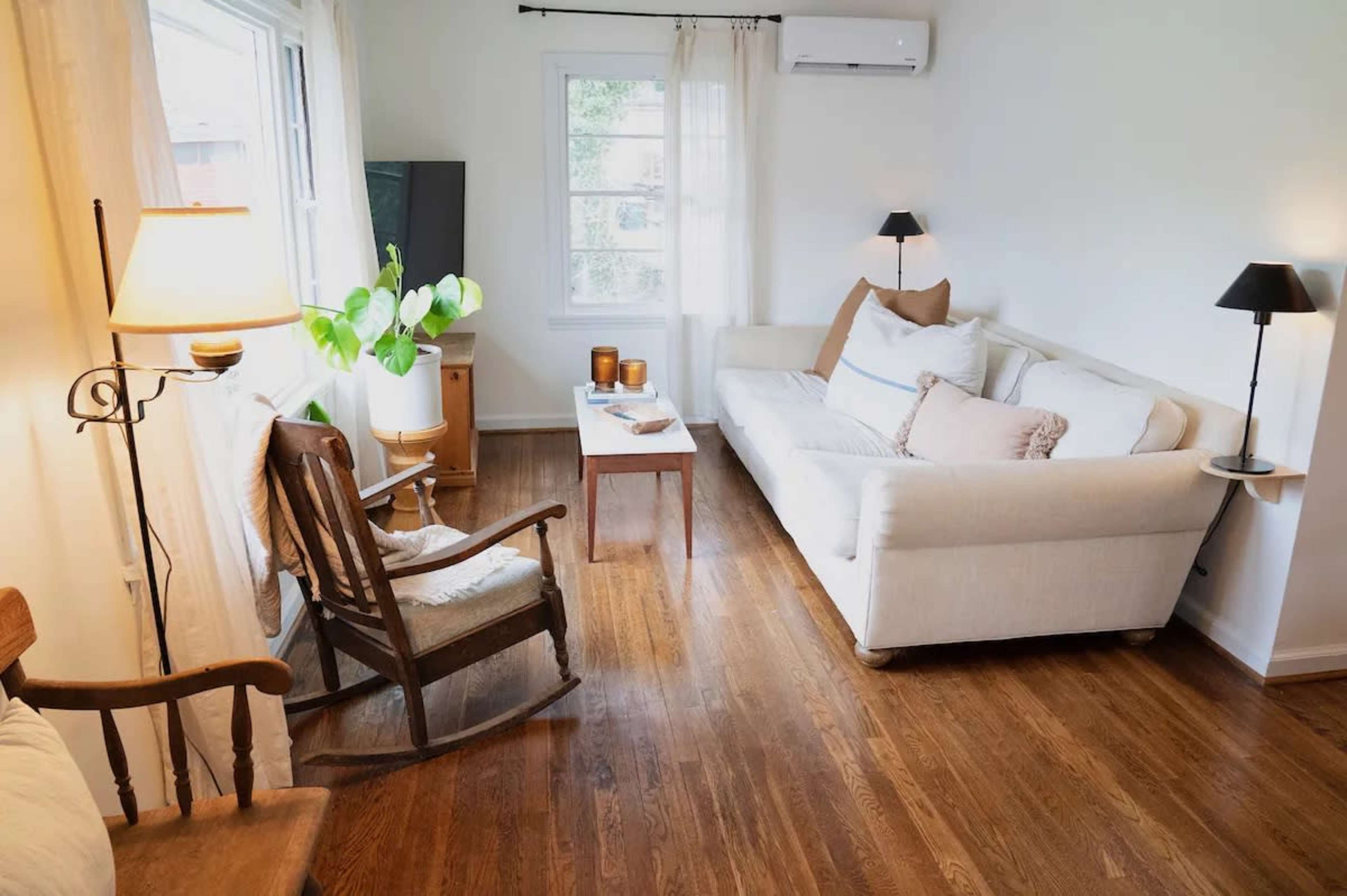The scene shows a living room with a white sofa, a small coffee table, and a rocking chair, all arranged around a large window allowing natural light to enter.