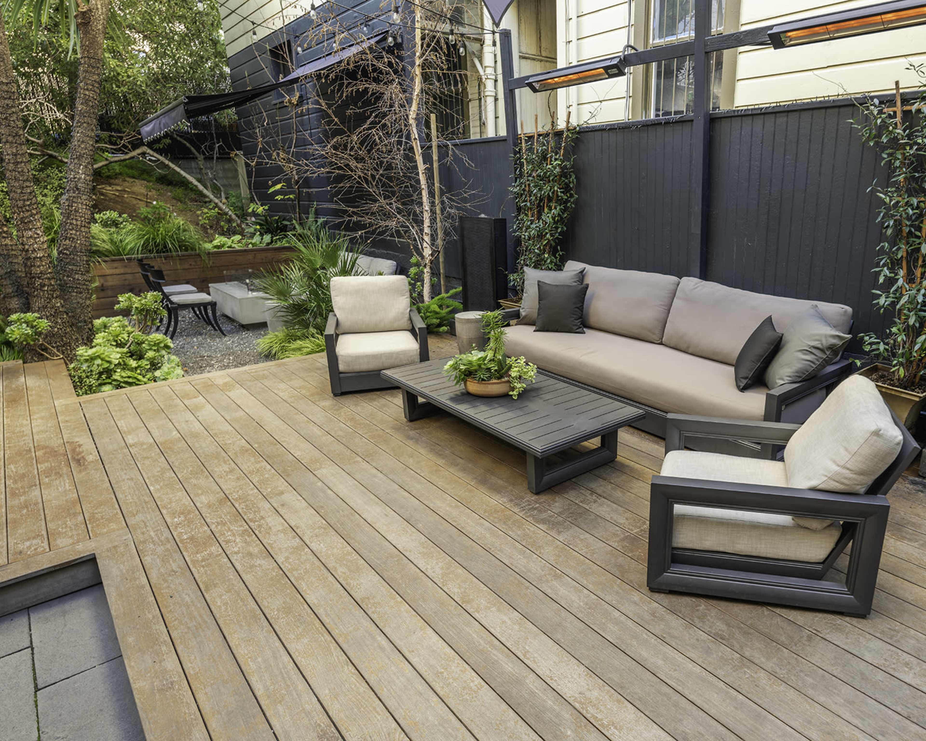The image shows a wooden deck with a grey outdoor sofa, two armchairs, and a coffee table, surrounded by greenery and a pathway in a landscaped backyard.