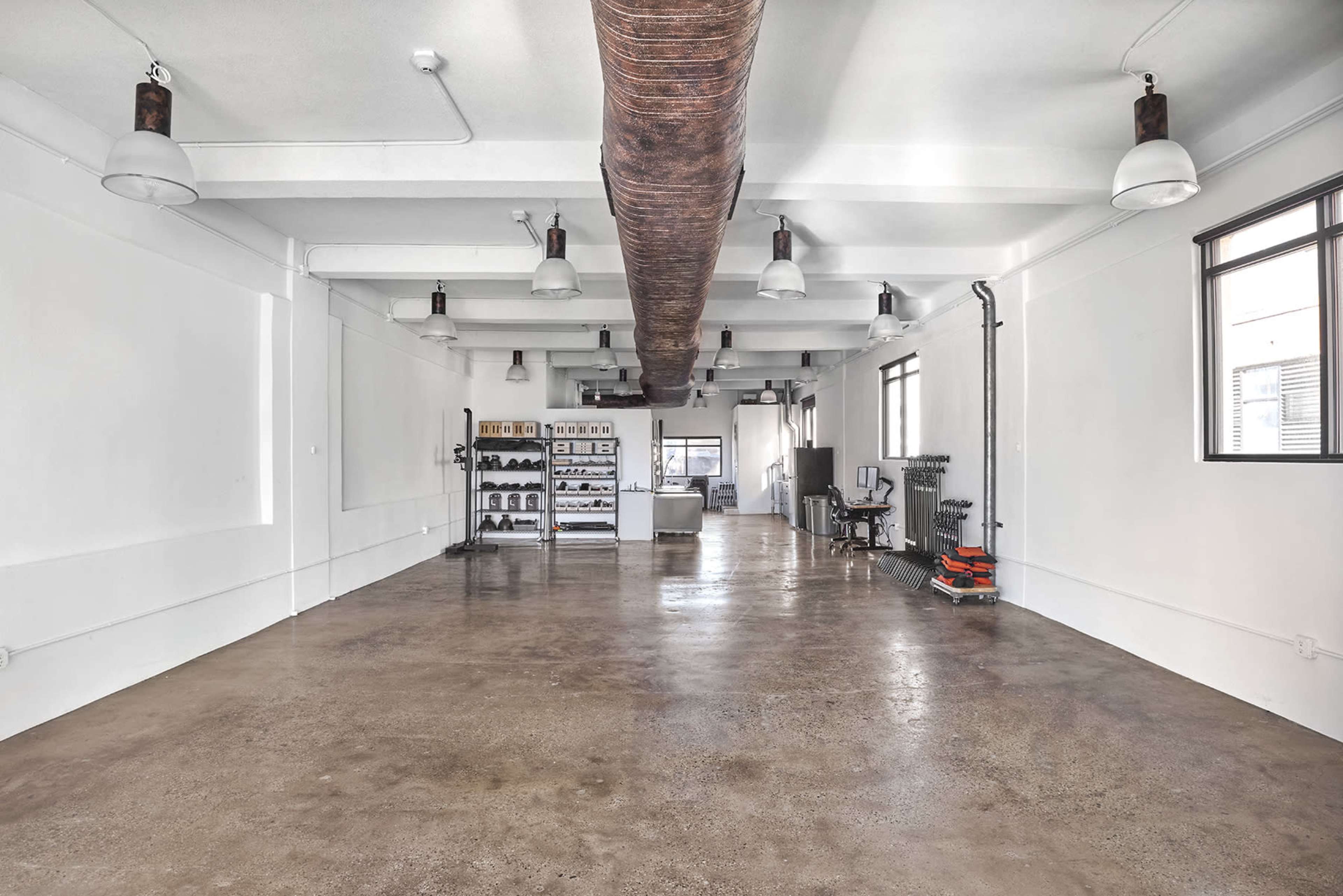 The image shows a spacious, minimalistic industrial interior with polished concrete floors, exposed pipes, and overhead lighting.