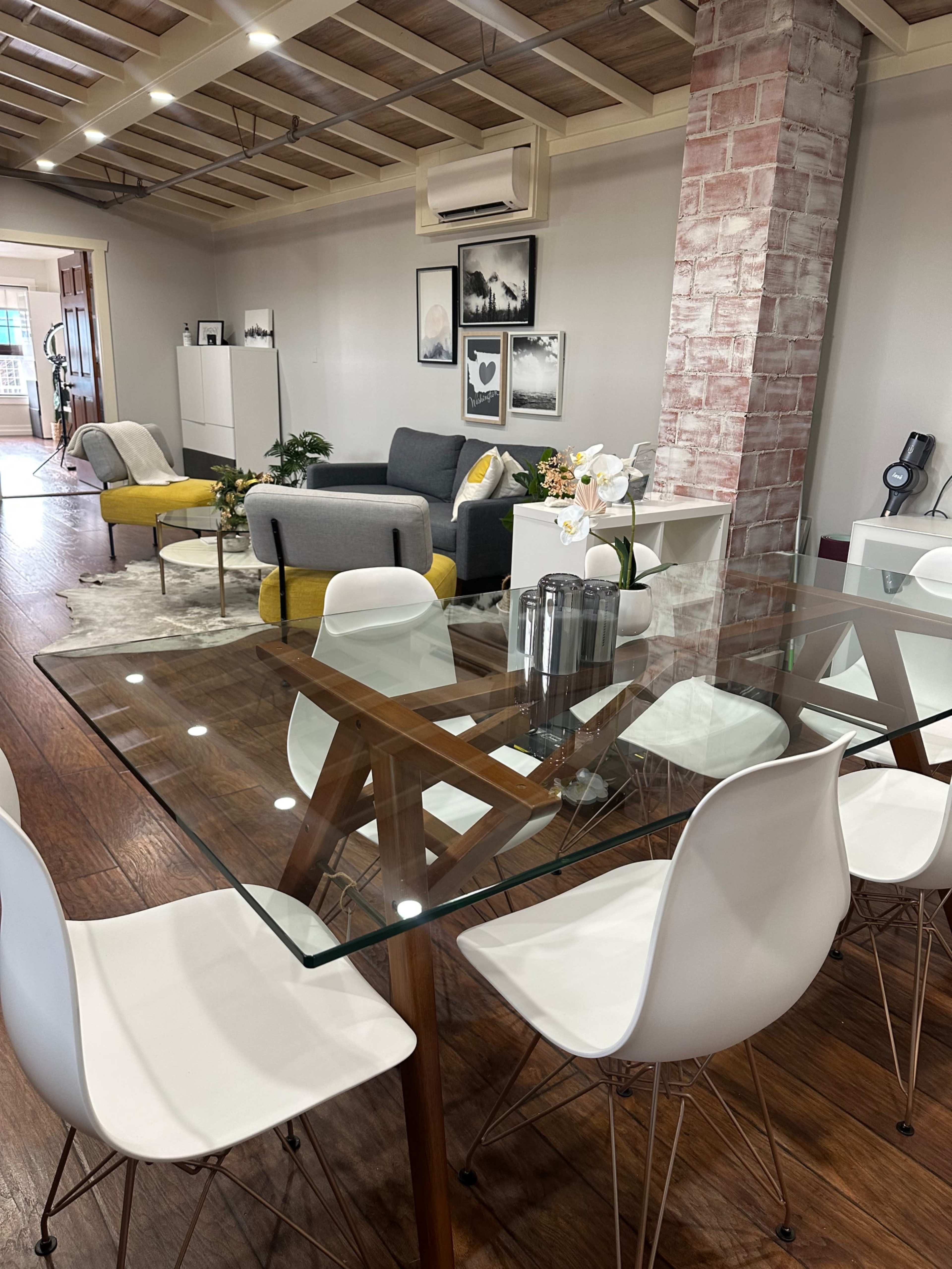 The image shows a modern living space featuring a glass dining table surrounded by white chairs, with a cozy seating area beyond that includes a gray sofa and decorative plants.
