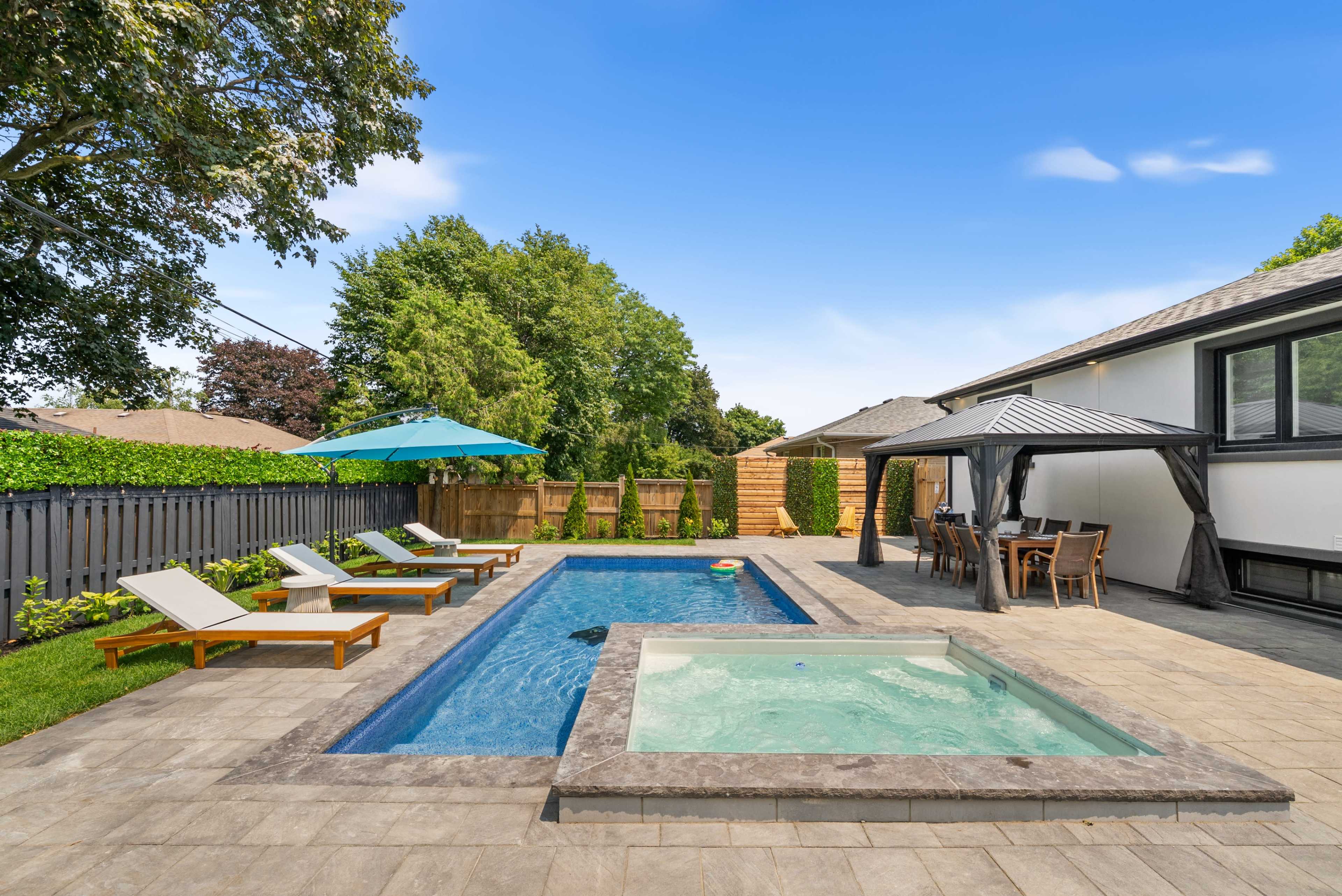 The scene features a backyard with a pool, a hot tub, lounge chairs, an umbrella, and a dining area under a gazebo.