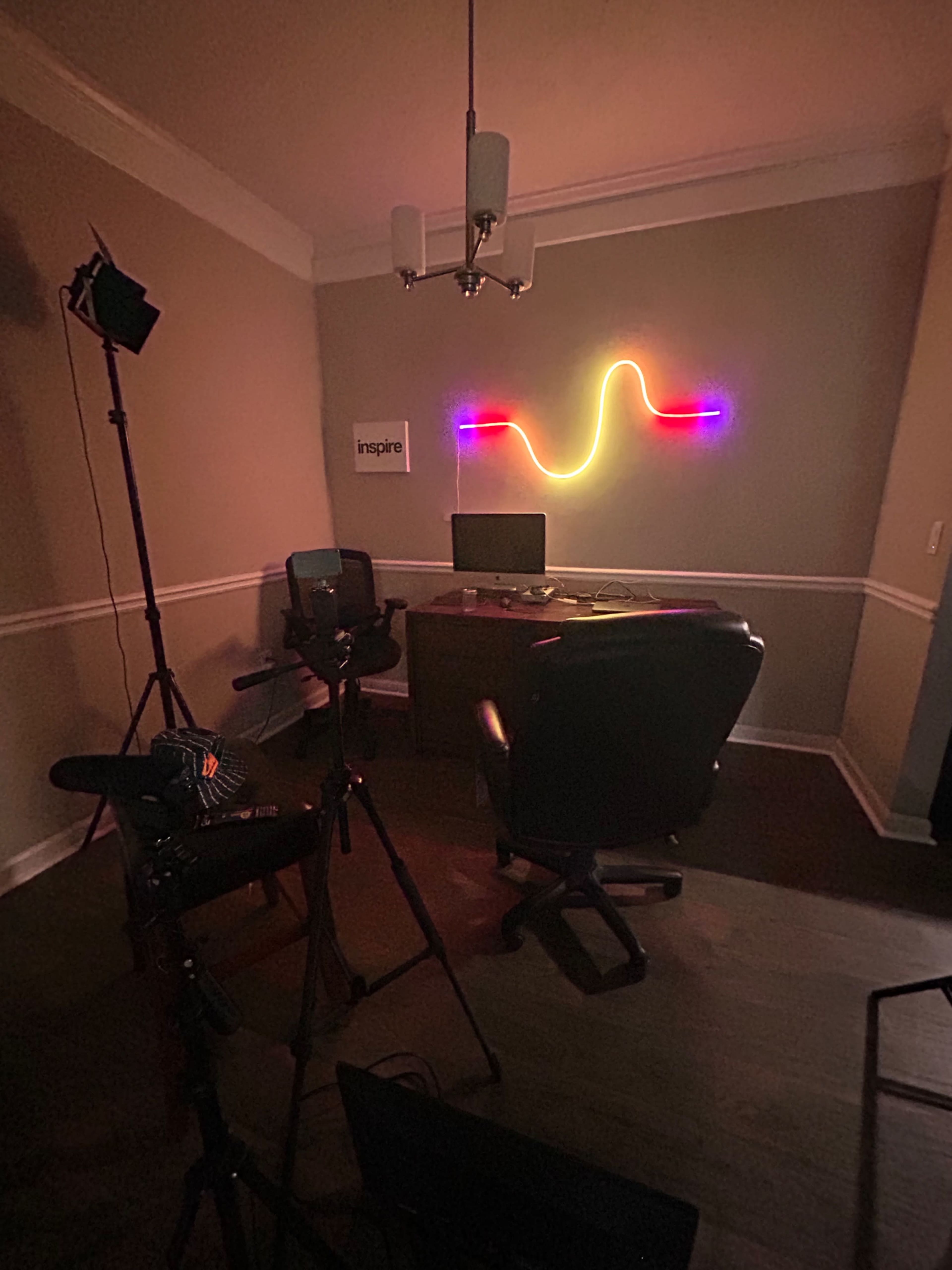 The image shows a dimly lit office space with a desk and an illuminated neon sign on the wall, along with photography equipment and a chair positioned facing the desk.