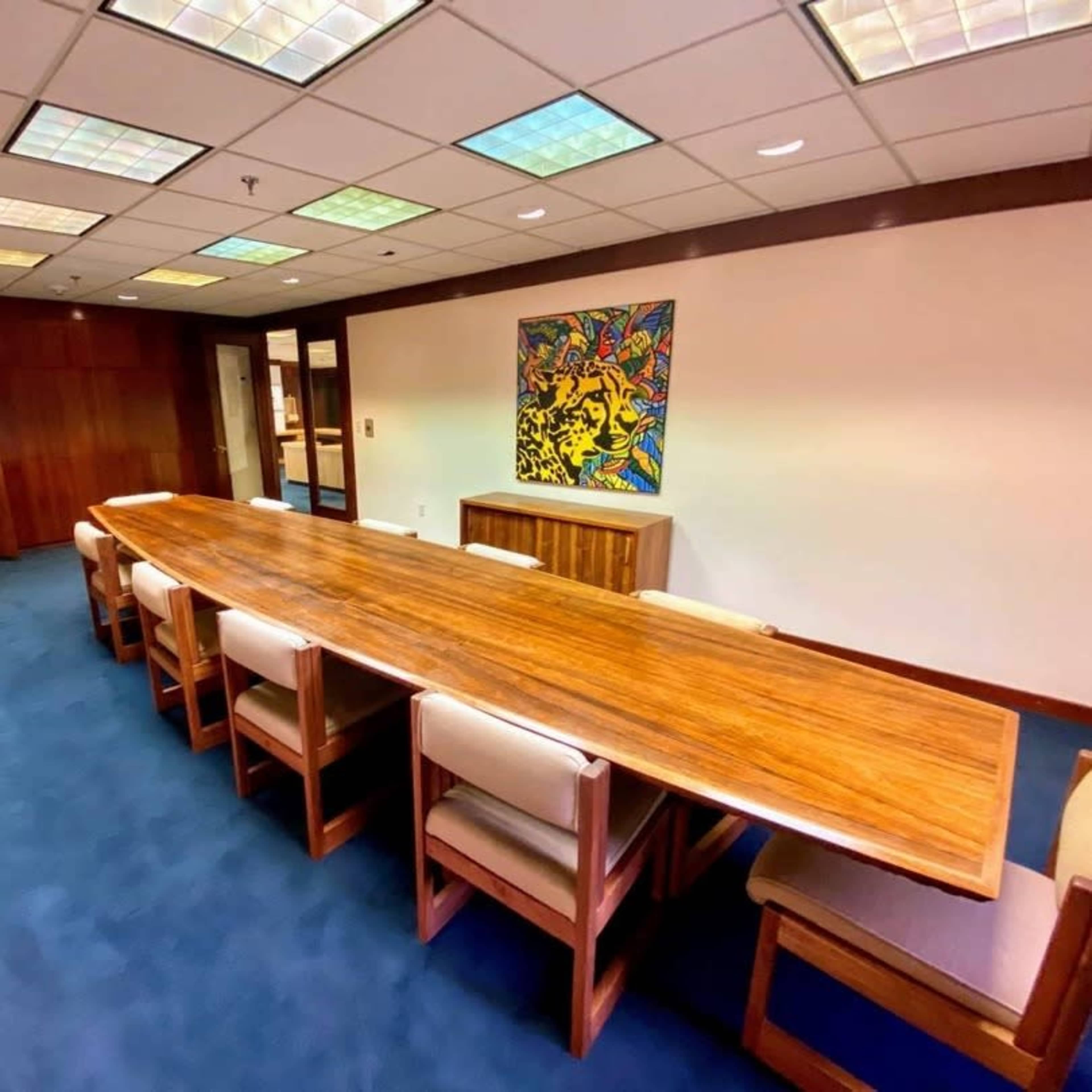 A long wooden conference table with eight chairs is positioned in a well-lit meeting room, featuring a colorful artwork on the wall.