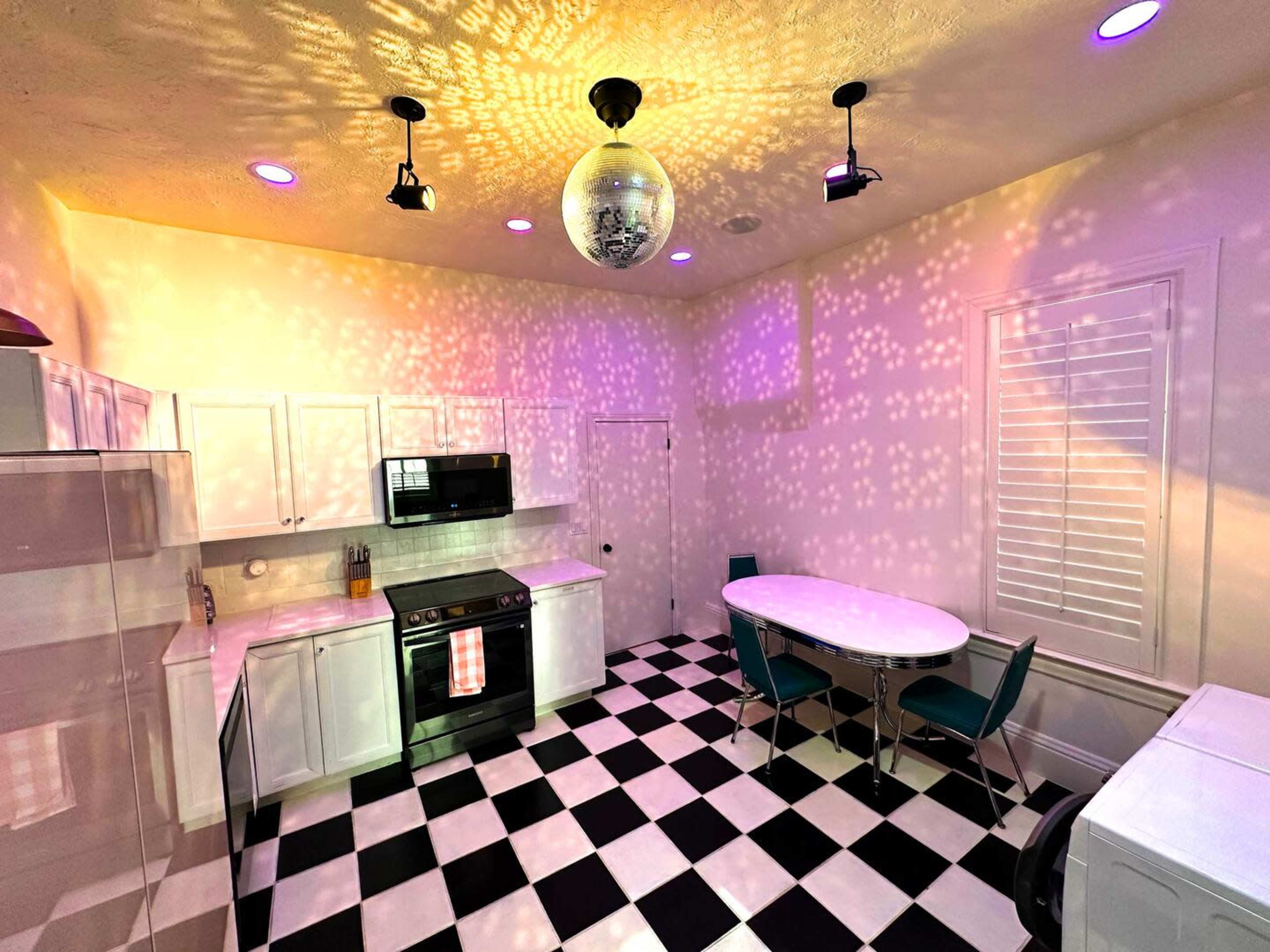 A modern kitchen with black and white checkered flooring, white cabinets, and colorful light reflections on the walls from a disco ball.