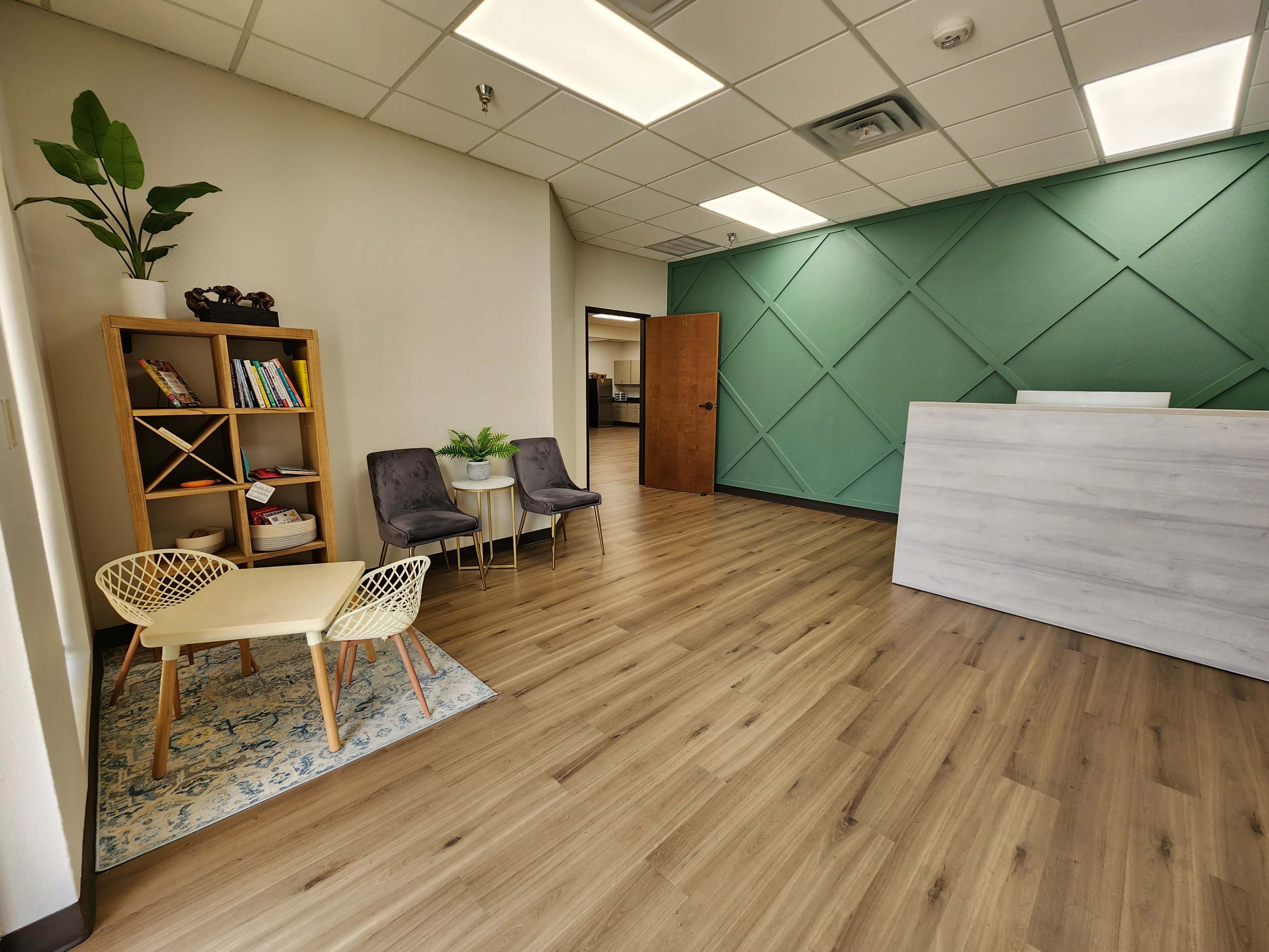 The image shows a modern office reception area with a green accent wall, a wooden bookshelf, two chairs, and a small table.
