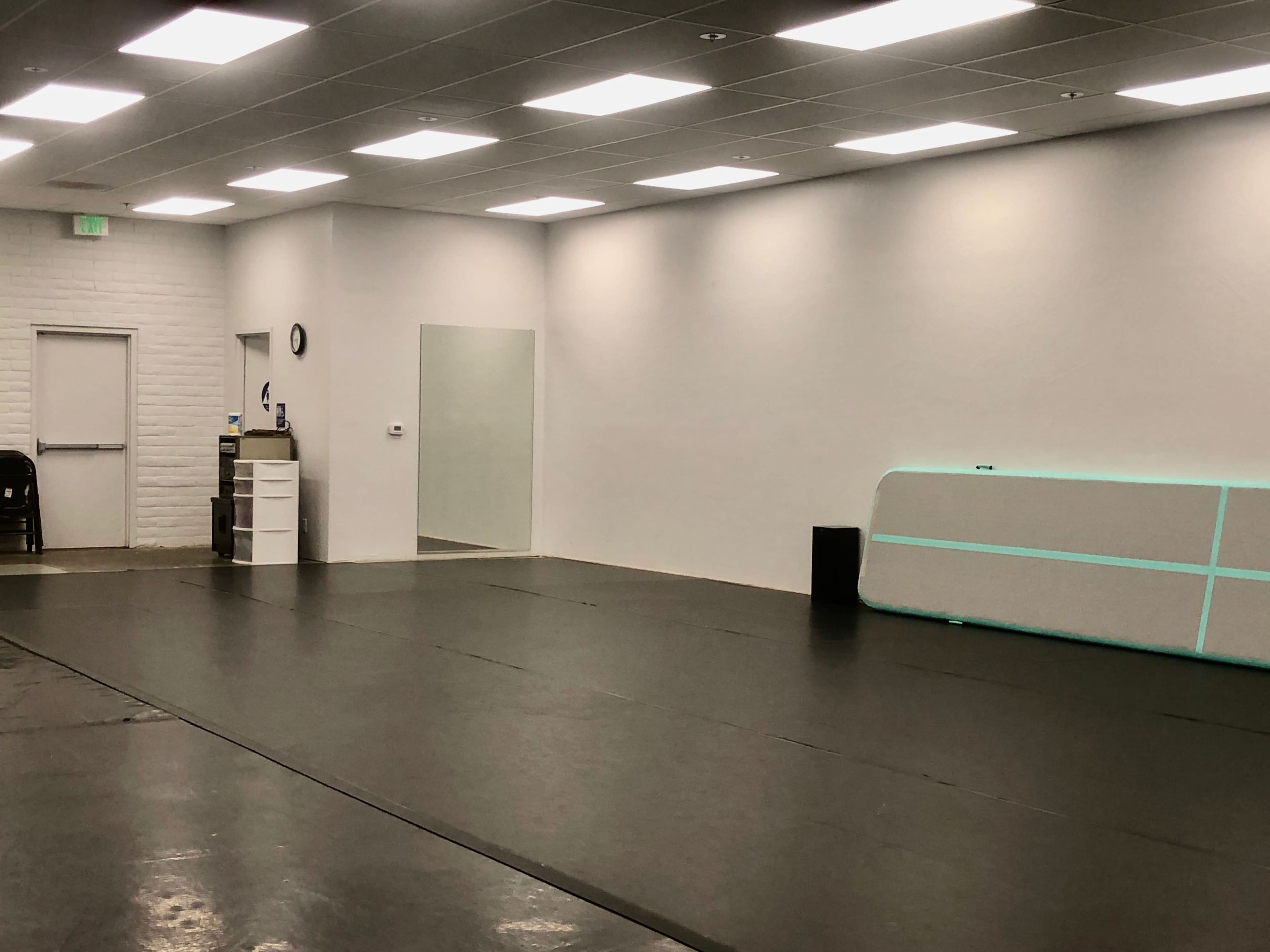 The image shows a spacious, empty room with a black floor, white walls, and bright overhead lights, featuring a mirror and a large foam wedge against the wall.