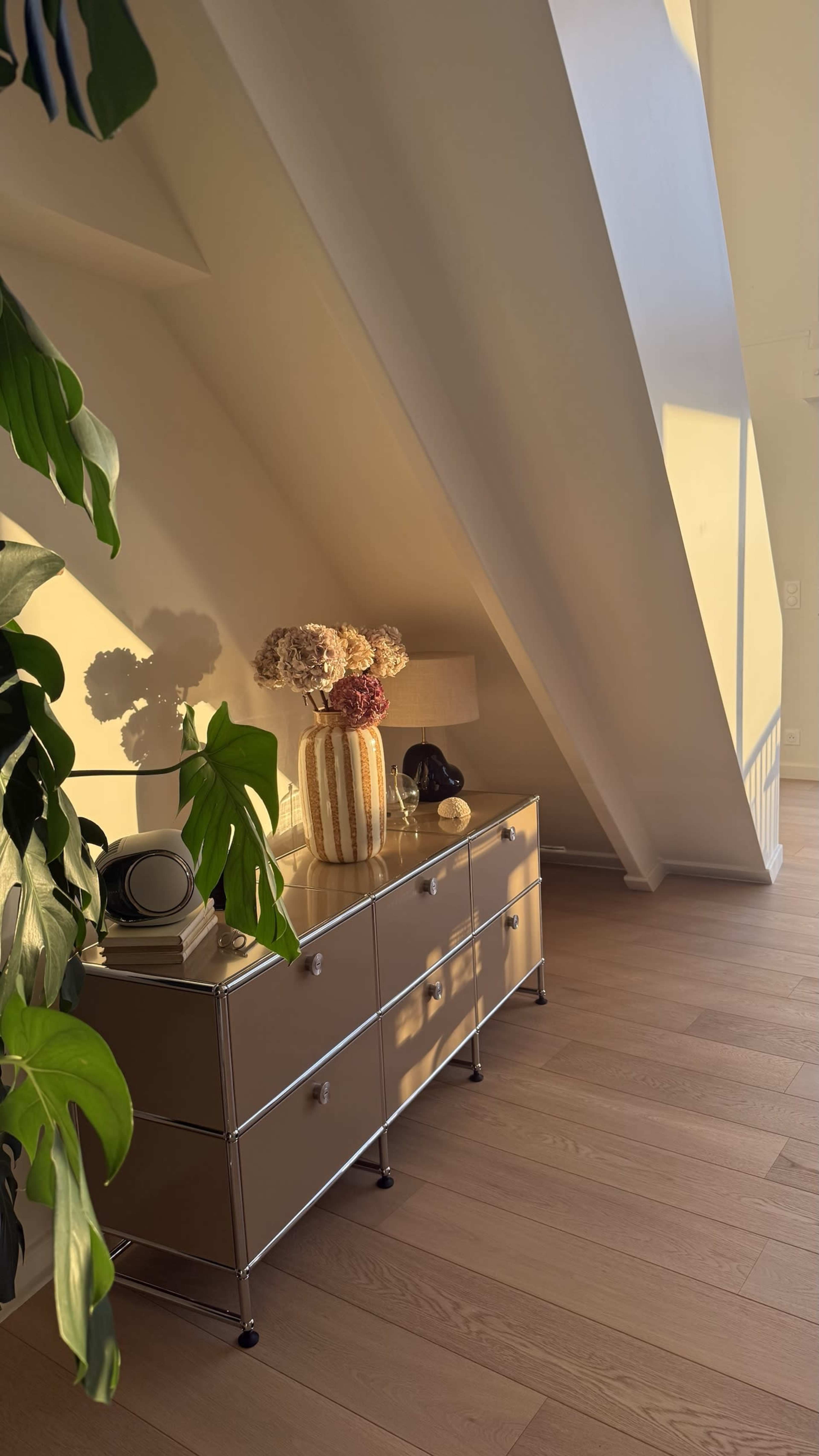 A dresser with a vase of dried flowers and a lamp is positioned against a slanted wall, with sunlight streaming in and casting shadows on the floor.