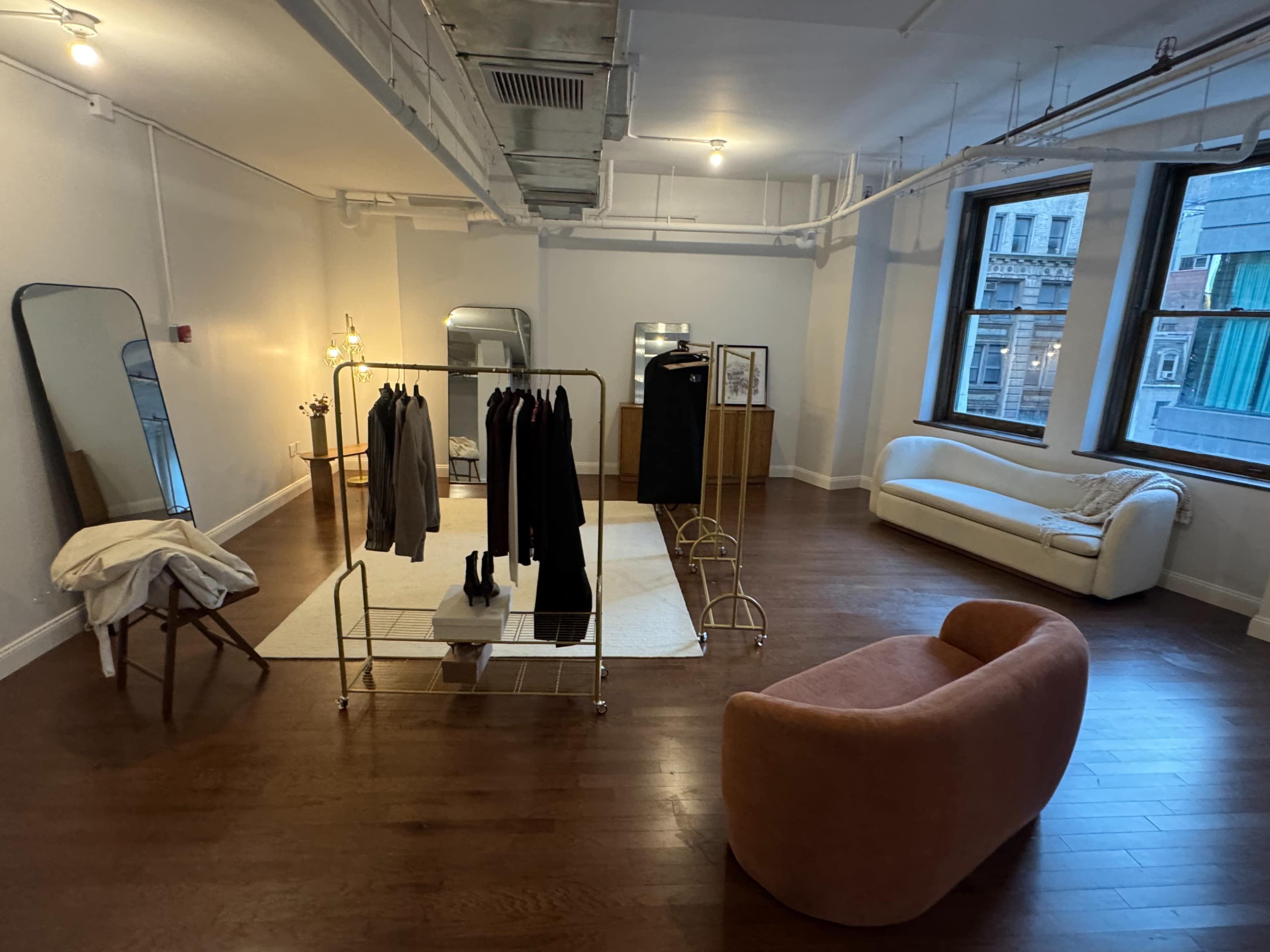 The image shows a spacious, well-lit interior featuring a clothing rack with garments, a large mirror, a minimalist sofa, and a wooden dresser in a contemporary style.