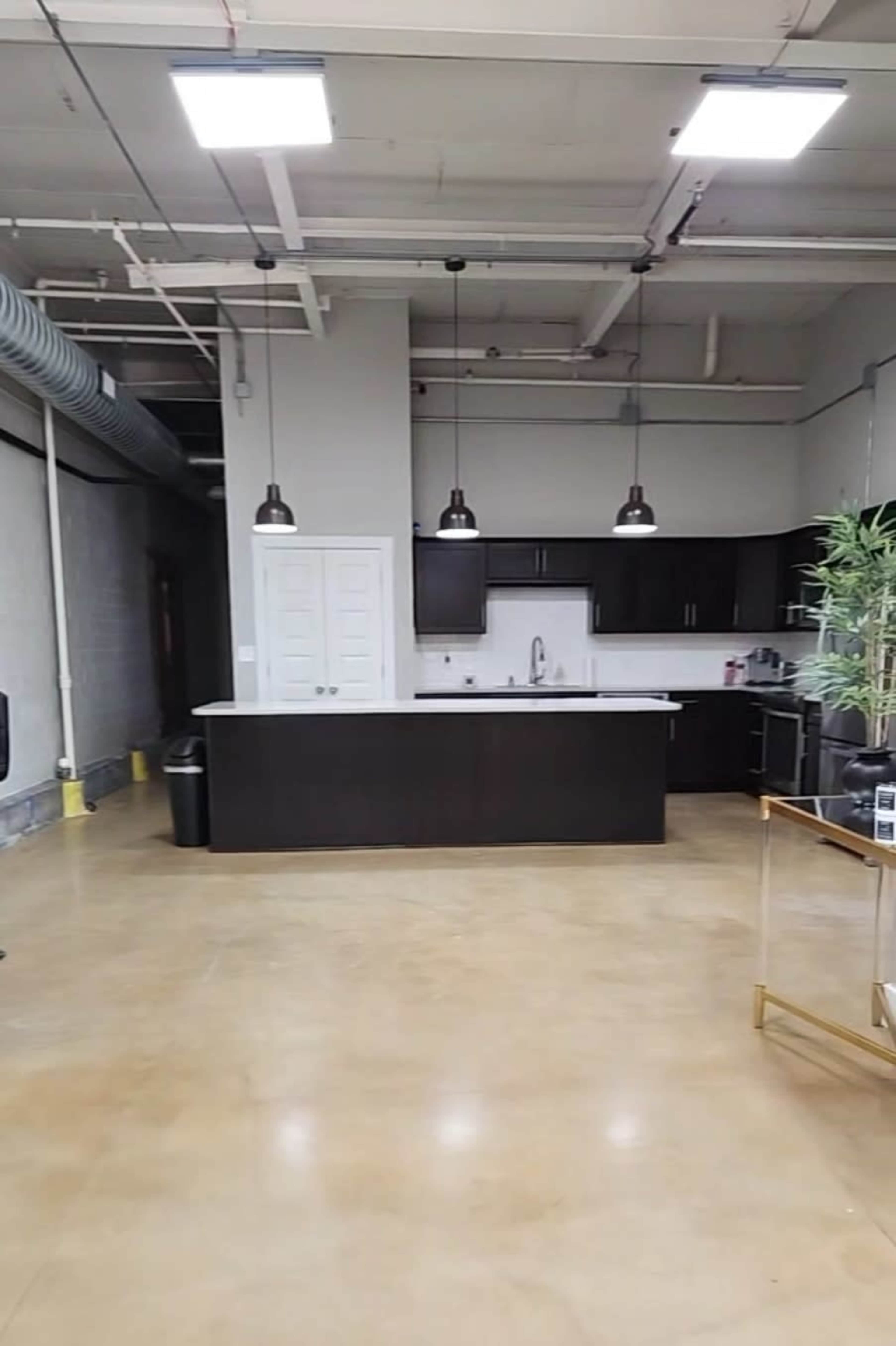 A modern kitchen area with a dark cabinet setup, a white countertop, and industrial-style lighting in an open office space.