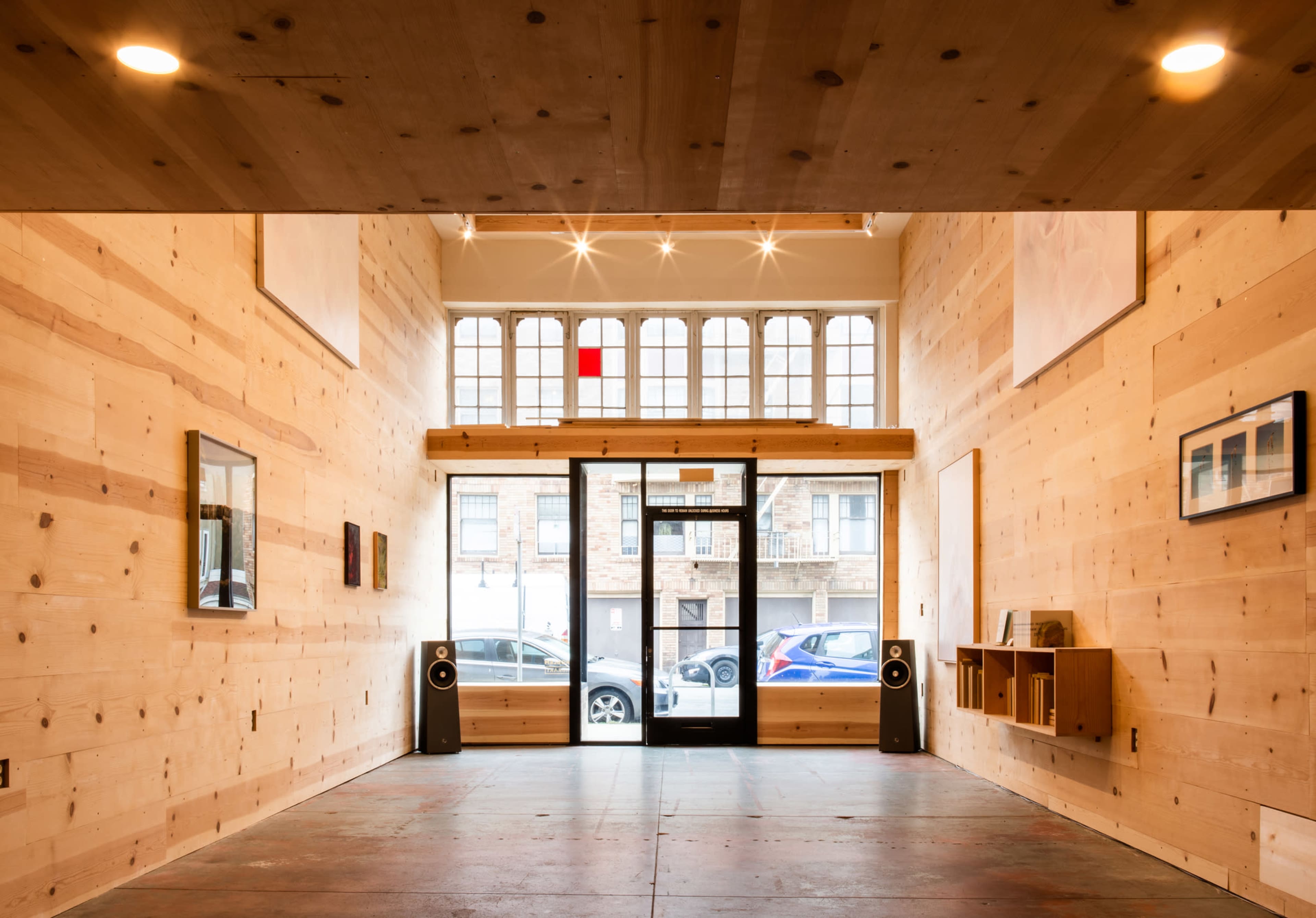 A wooden interior space with large windows, two speakers on the floor, and minimal furnishings along the walls.