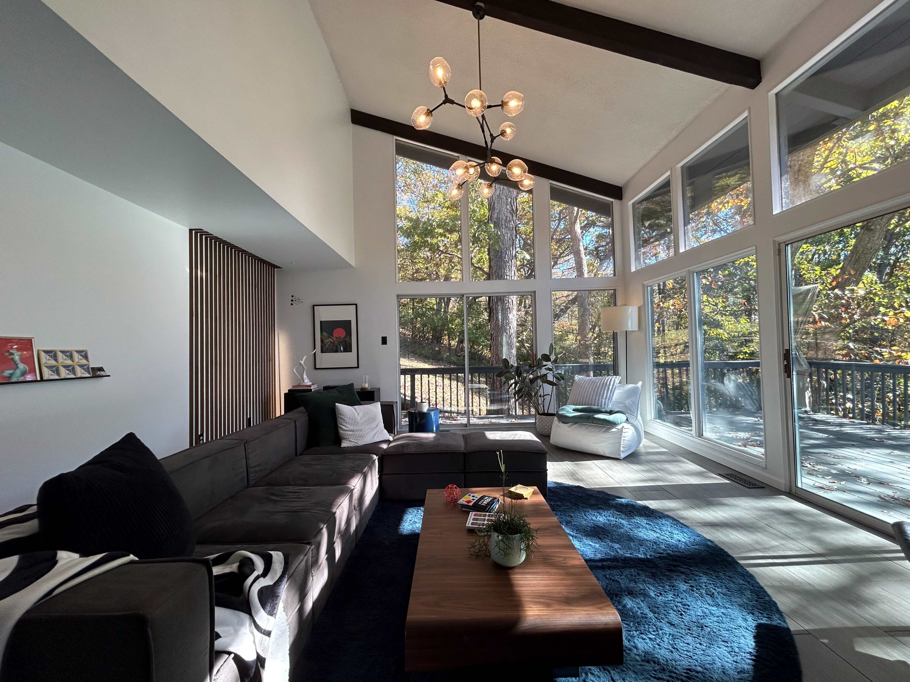 A modern living room features a large sectional sofa, a coffee table, and floor-to-ceiling windows that overlook a wooded area.