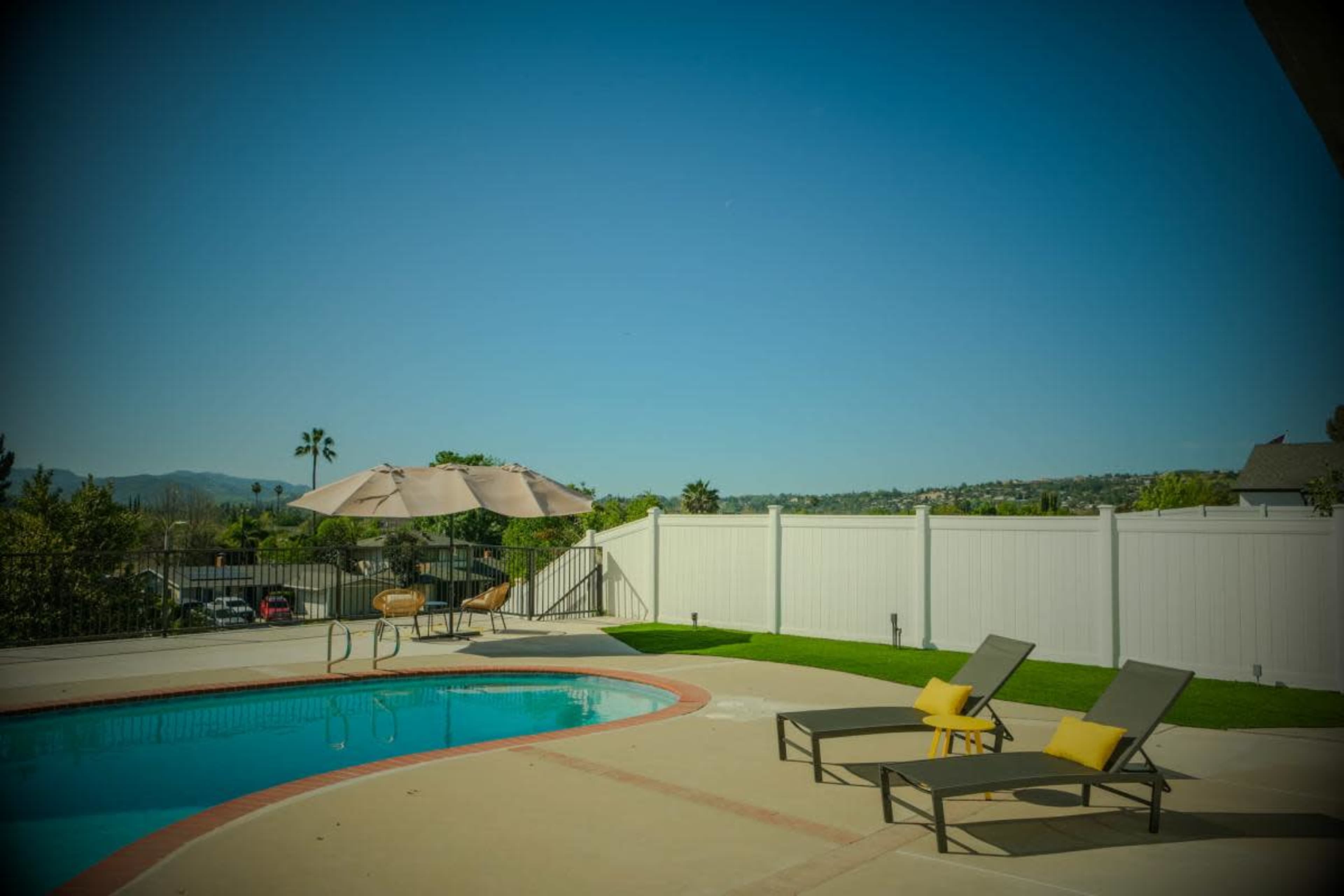 A pool area features two lounge chairs with yellow cushions, a table with chairs under an umbrella, and a view of a distant hillside.