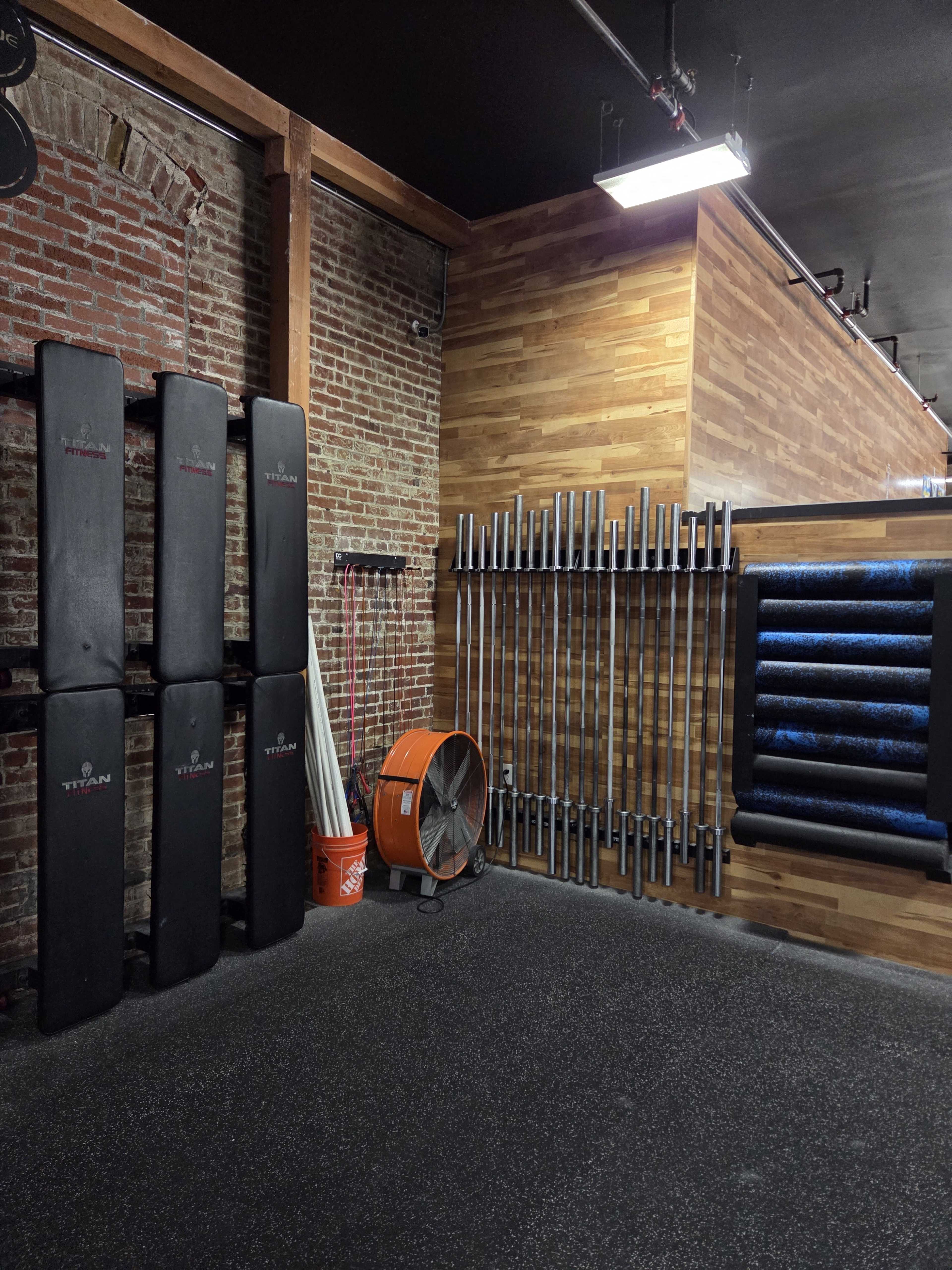 The image shows a gym corner with black workout mats stacked against a brick wall, several metal bars organized on a vertical rack, and a fan positioned beside a bucket.