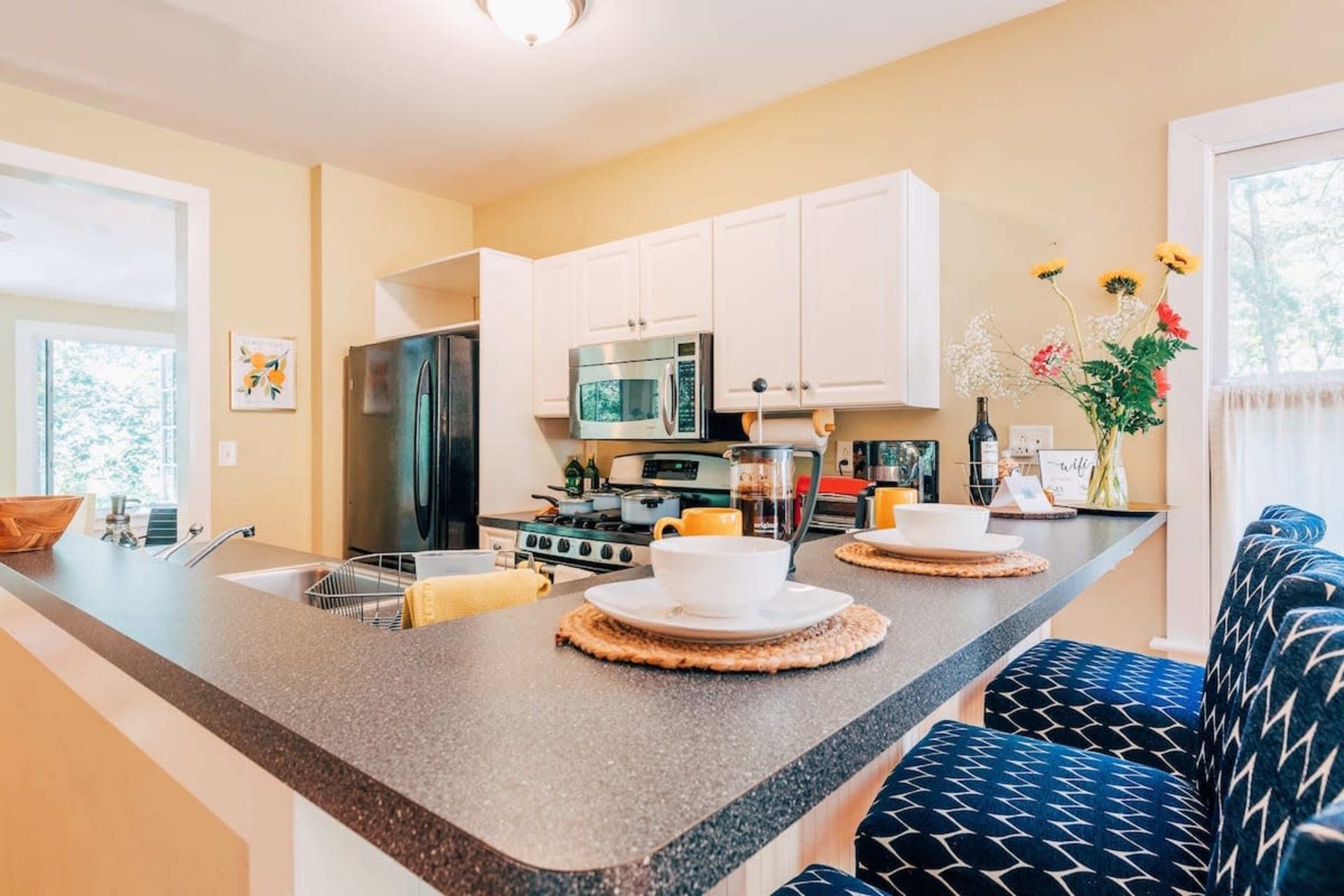 A modern kitchen layout features a countertop with two place settings, a stainless steel stove, and white cabinetry adorned with a vase of flowers.