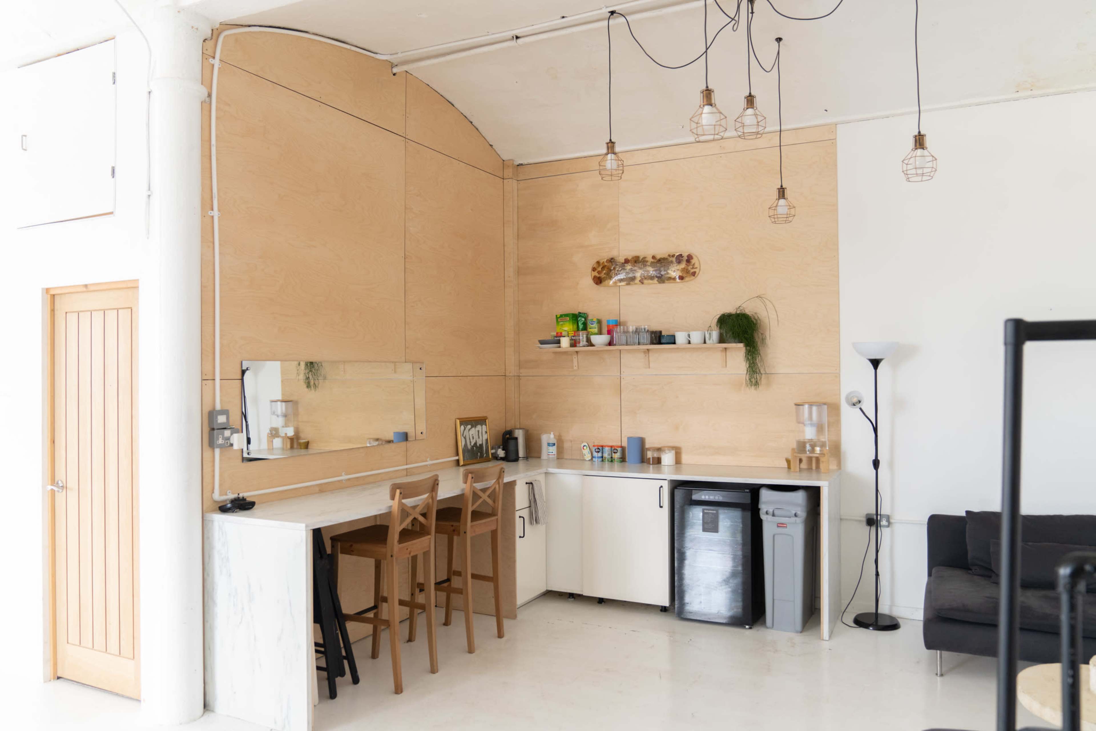 The image shows a minimalist kitchen area with wooden paneling, a countertop, three bar stools, and a shelf holding plants and kitchen items.