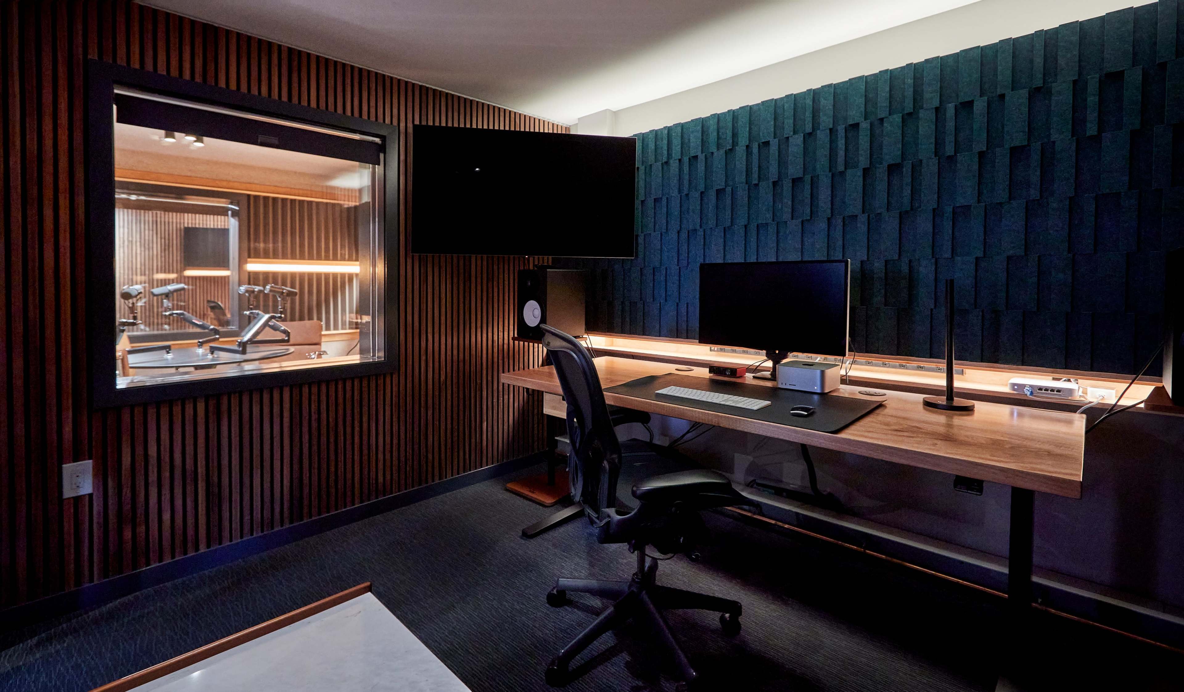 A modern recording studio with a dark wood panelled wall, a large desk with monitors, and a glass-enclosed sound booth visible in the background.