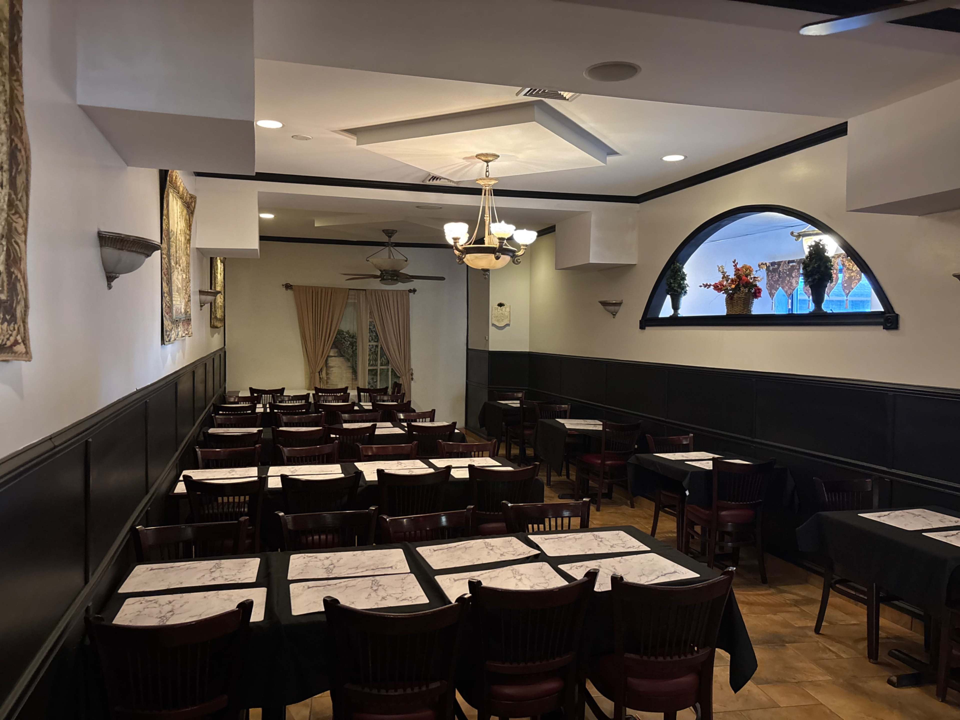 The image shows an empty dining area of a restaurant, featuring rows of tables covered with black cloths and arranged with chairs, illuminated by ceiling lights.