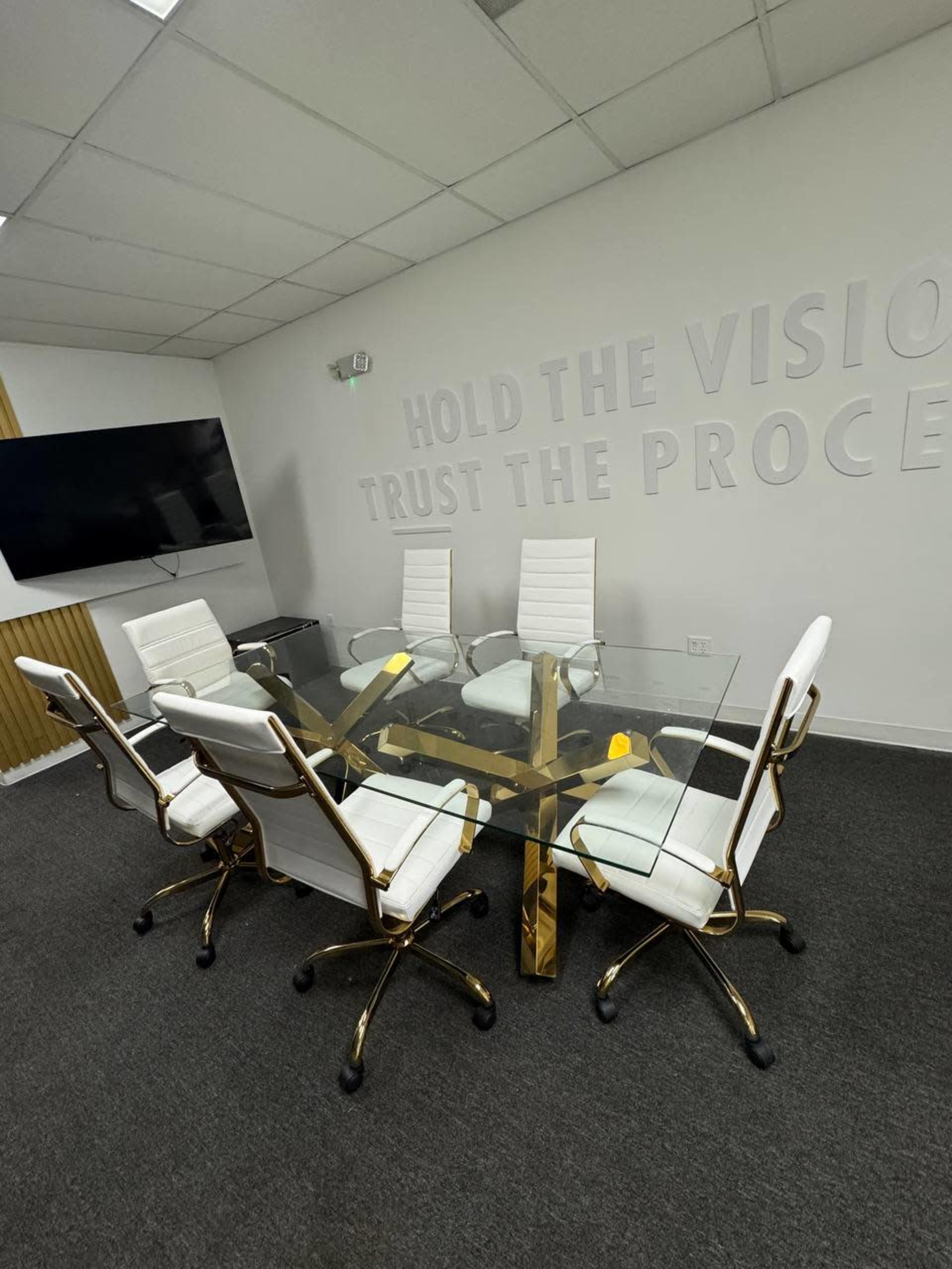 A modern conference room features a glass table surrounded by six white chairs, with a wall displaying the phrase "HOLD THE VISION TRUST THE PROCESS."