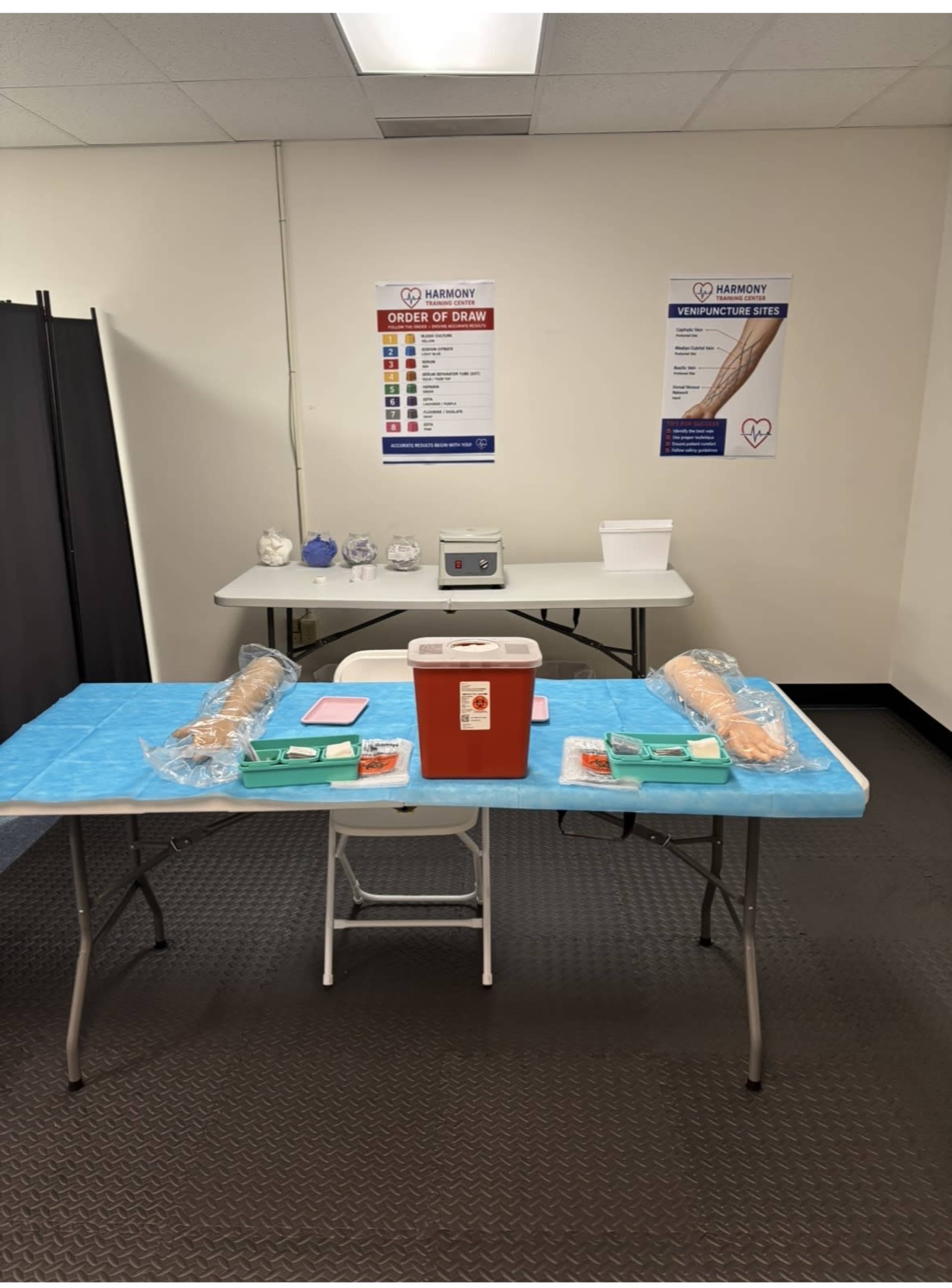 The image shows a room setup for a medical procedure, featuring a blue table with two long padded arms, a red cooler, and a table with medical supplies in the background.