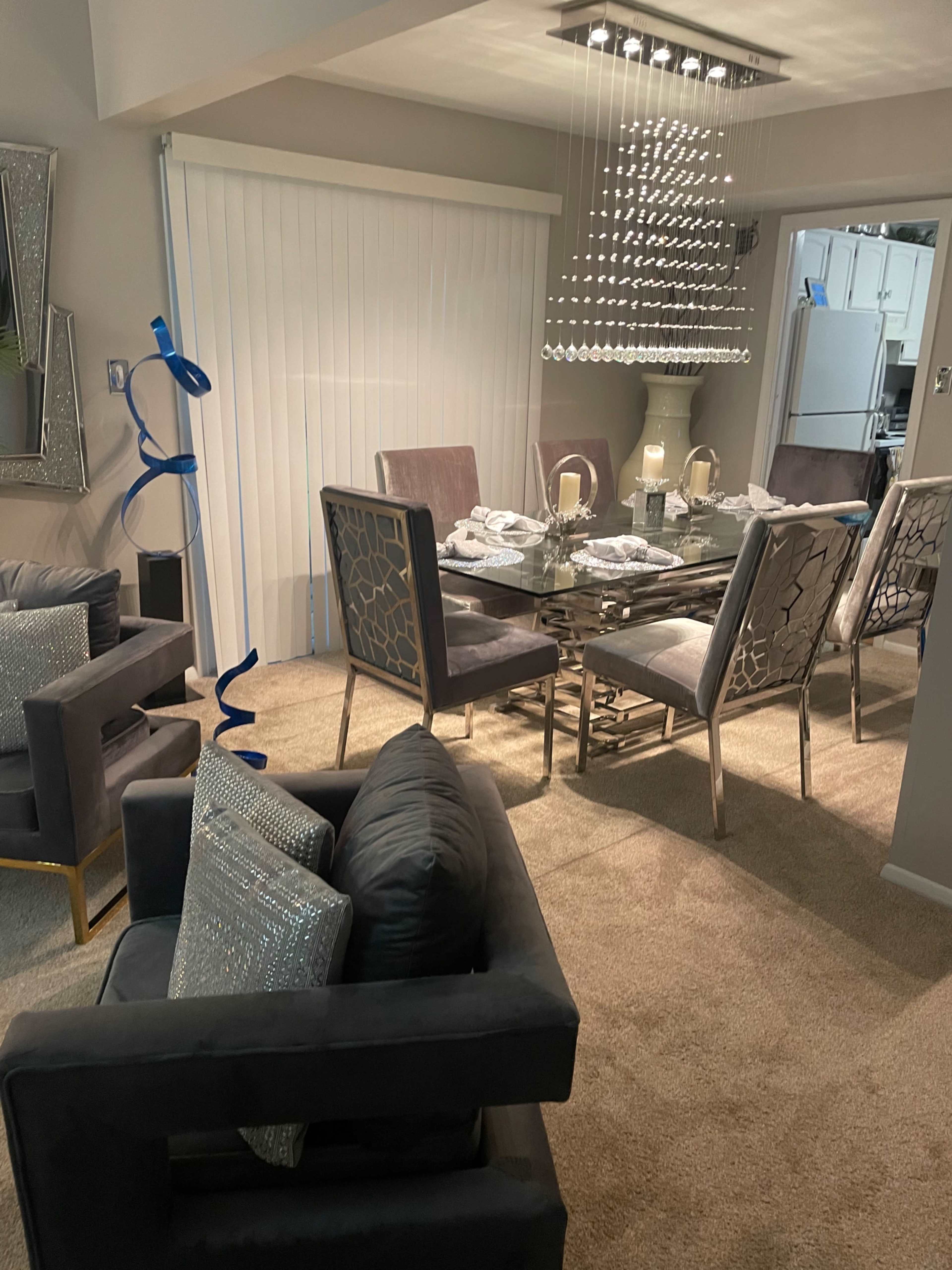 A dining area features a glass table set for six with modern chairs, surrounded by carpeted flooring and a large window covered by blinds.