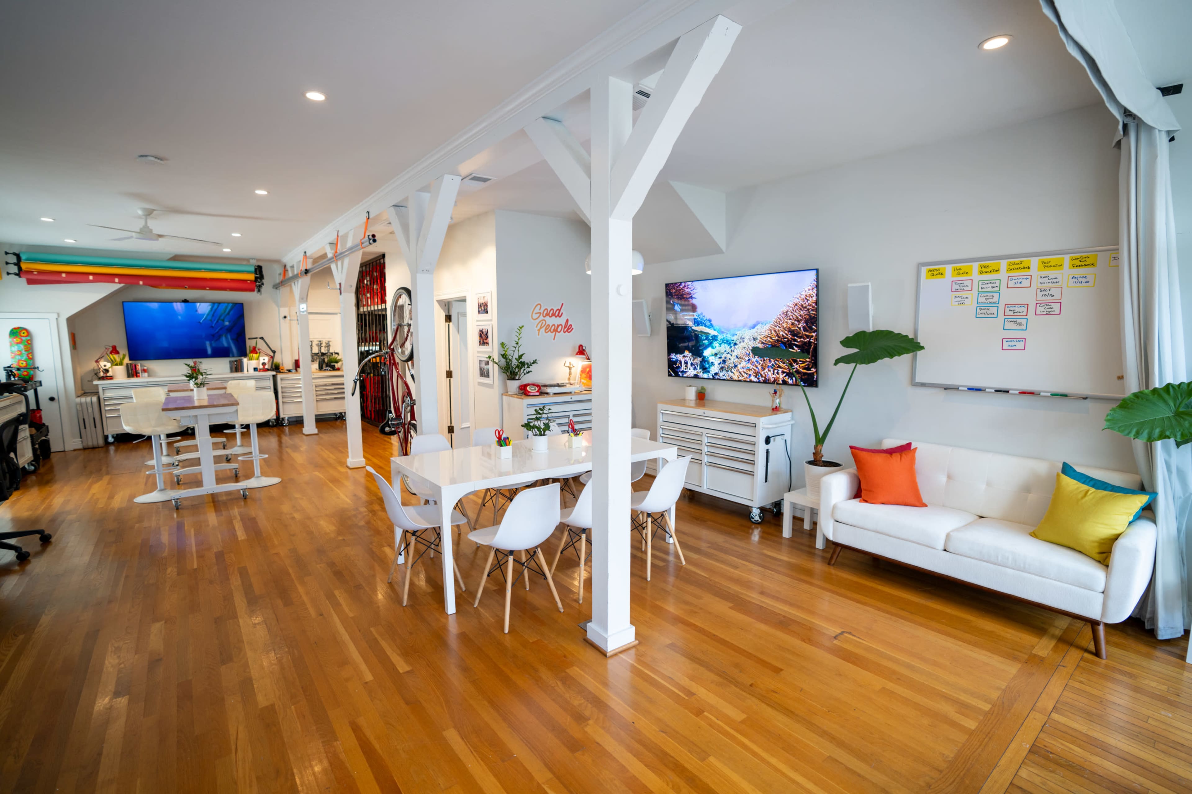 The image shows a modern open-concept living space with wooden flooring, a dining table, white chairs, a couch, and two large televisions mounted on the walls.