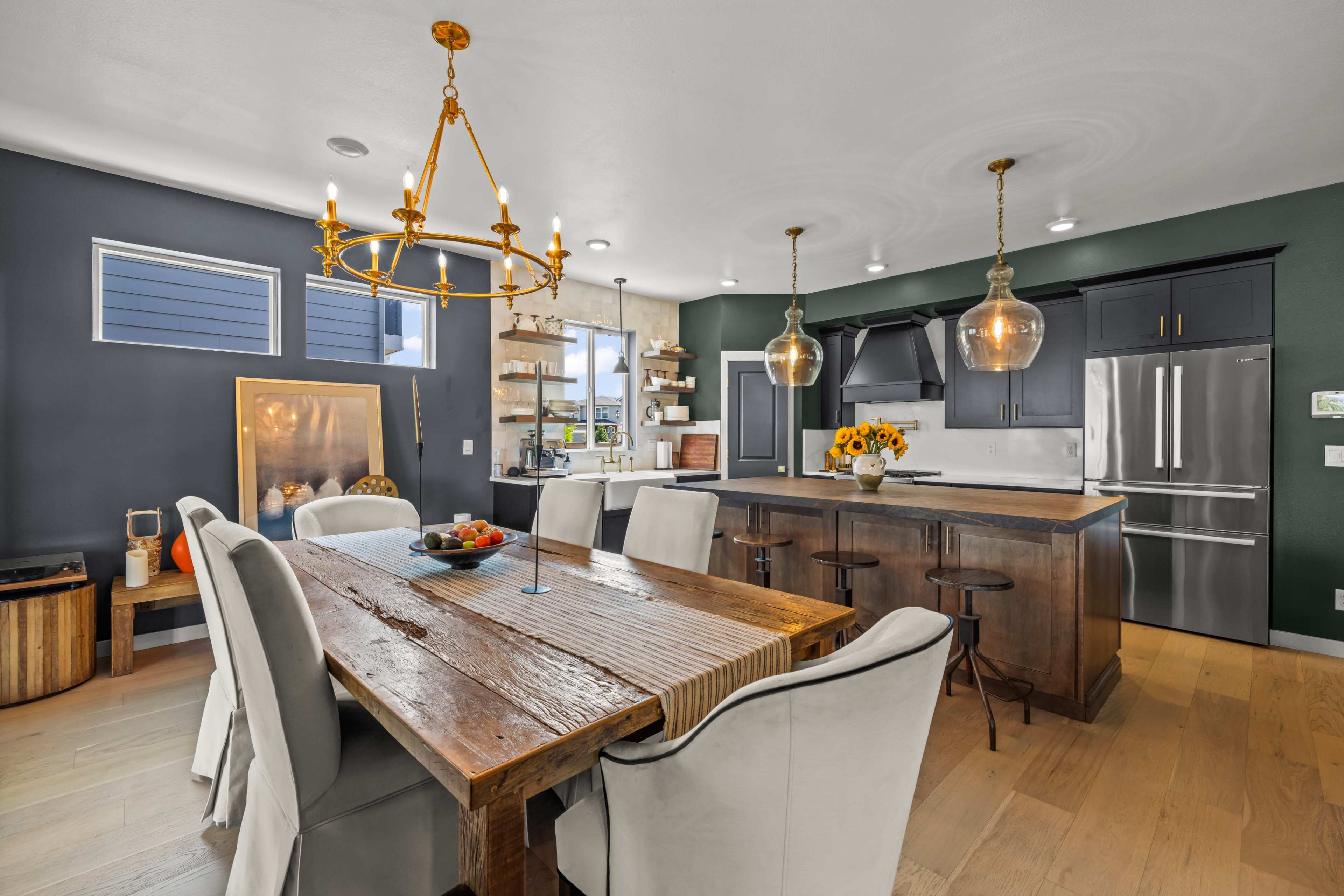 The image shows a modern kitchen and dining area featuring a large wooden table with upholstered chairs, a kitchen island with pendant lighting, and a dark accent wall.