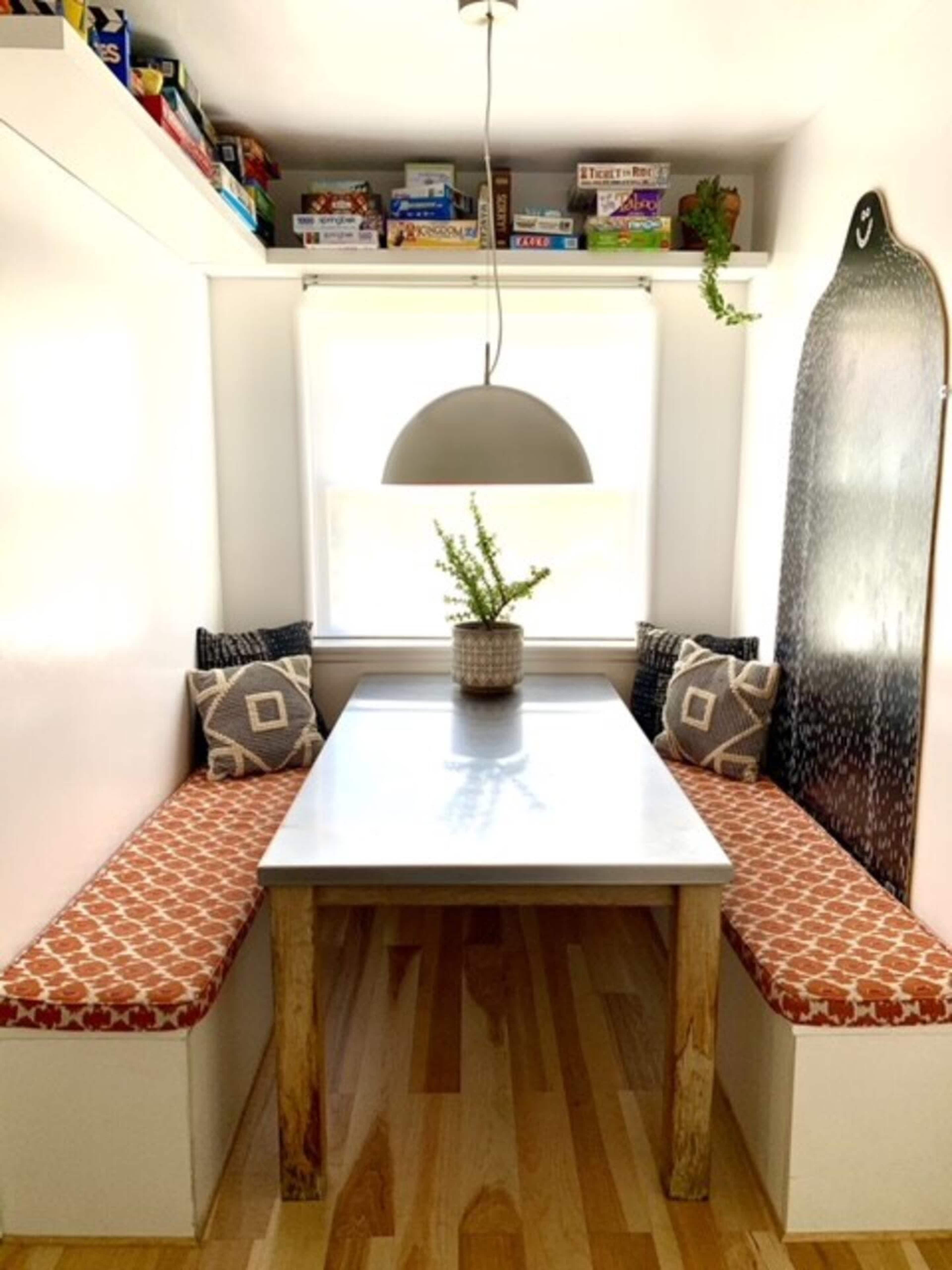 A small dining nook features a table surrounded by two upholstered benches and a large pendant light above, with shelves filled with board games in the background.