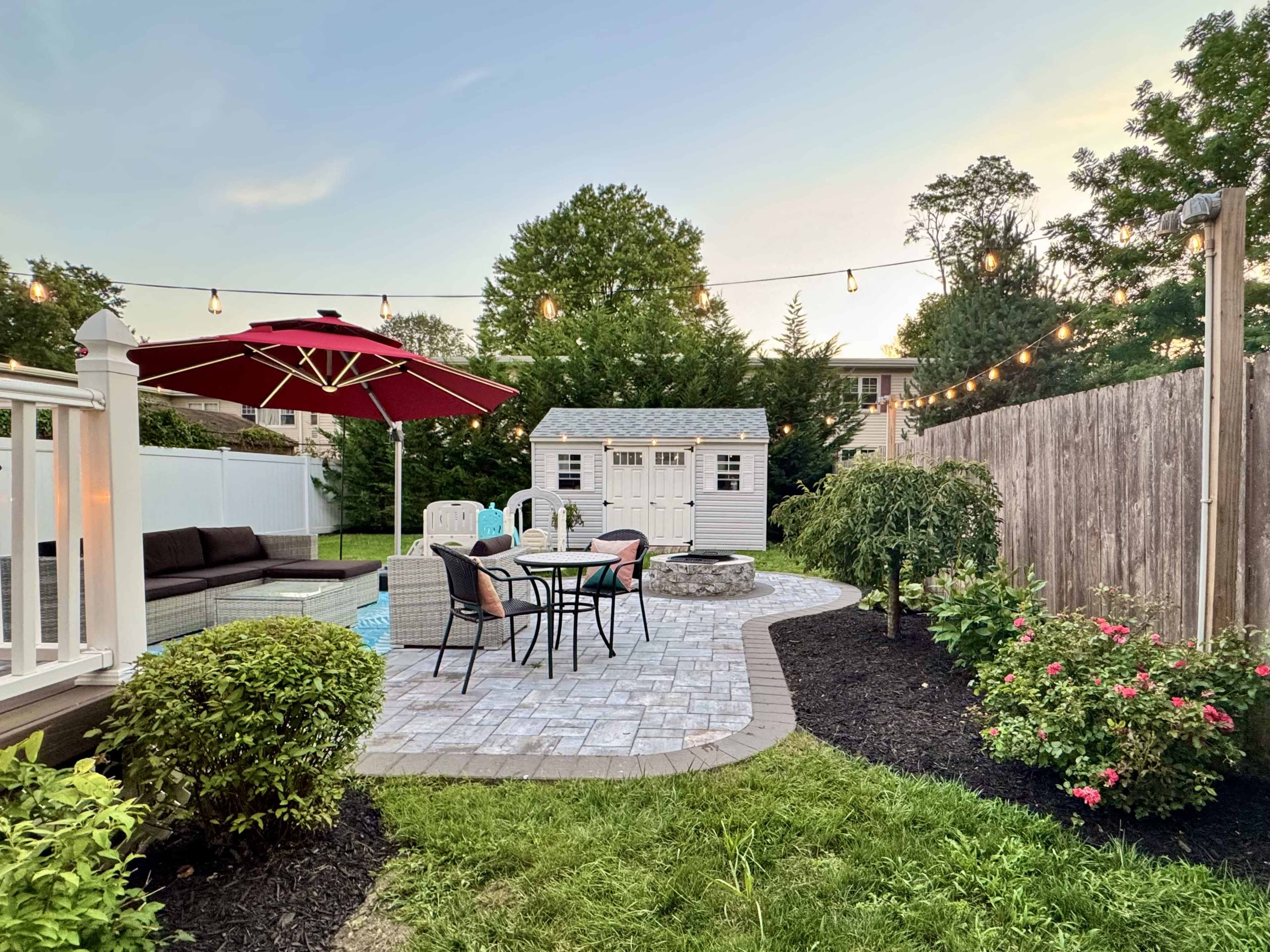 The image shows a landscaped backyard with a patio area, a shed, outdoor seating, and decorative lights strung overhead.