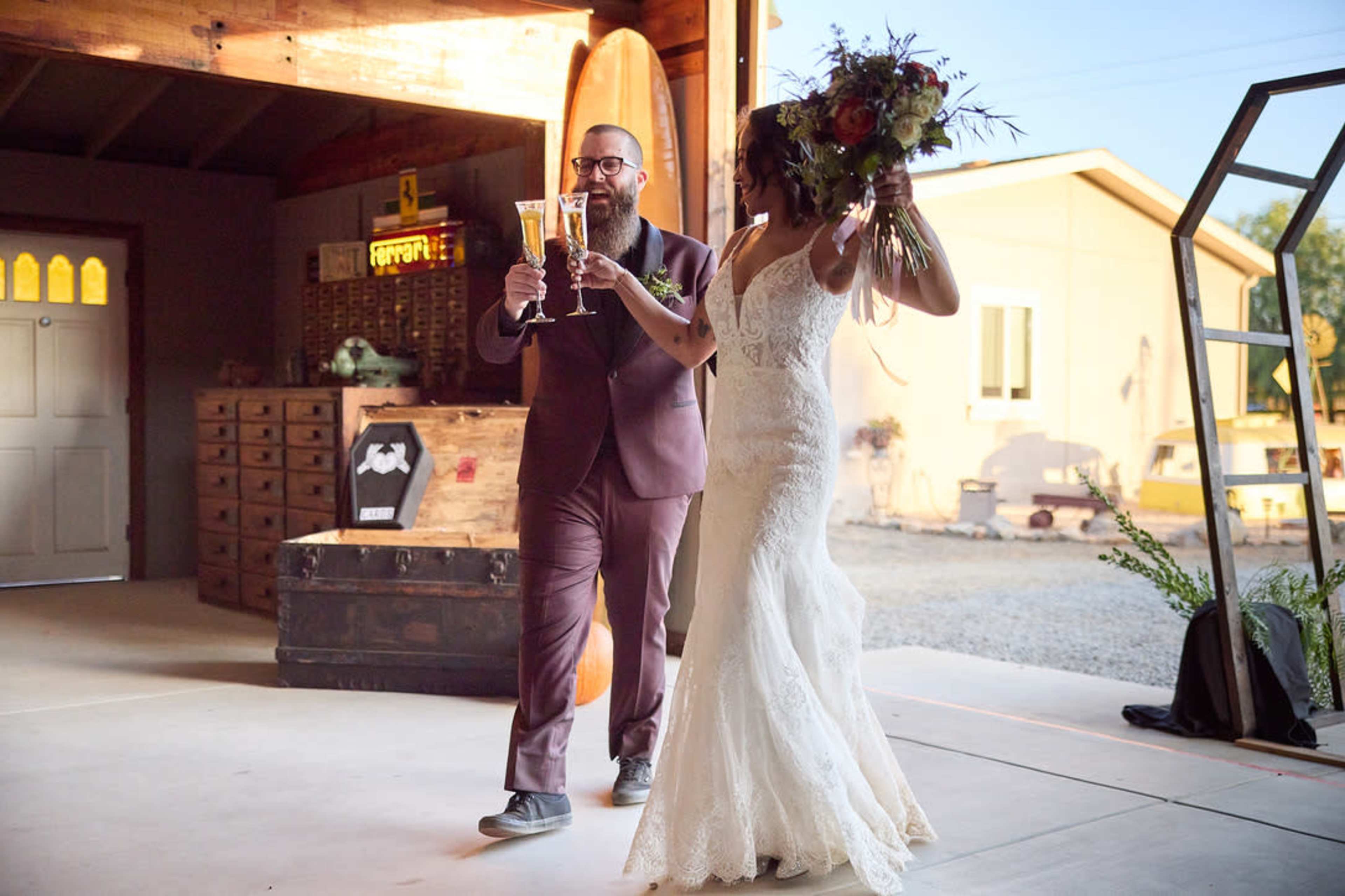 A man in a burgundy suit and a woman in a lace wedding dress enter a venue together, both holding drinks and smiling.