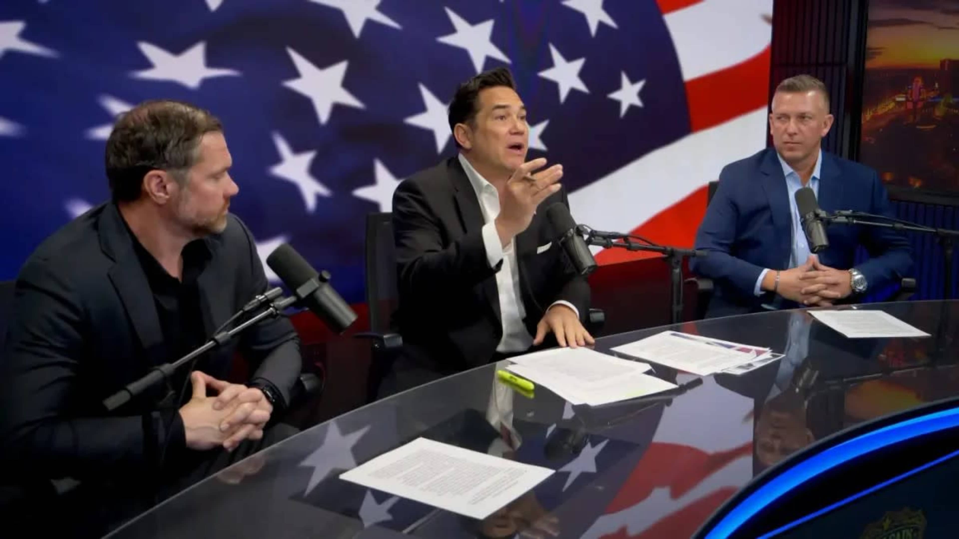 Three men sit at a table with microphones, discussing in front of a backdrop of the American flag.