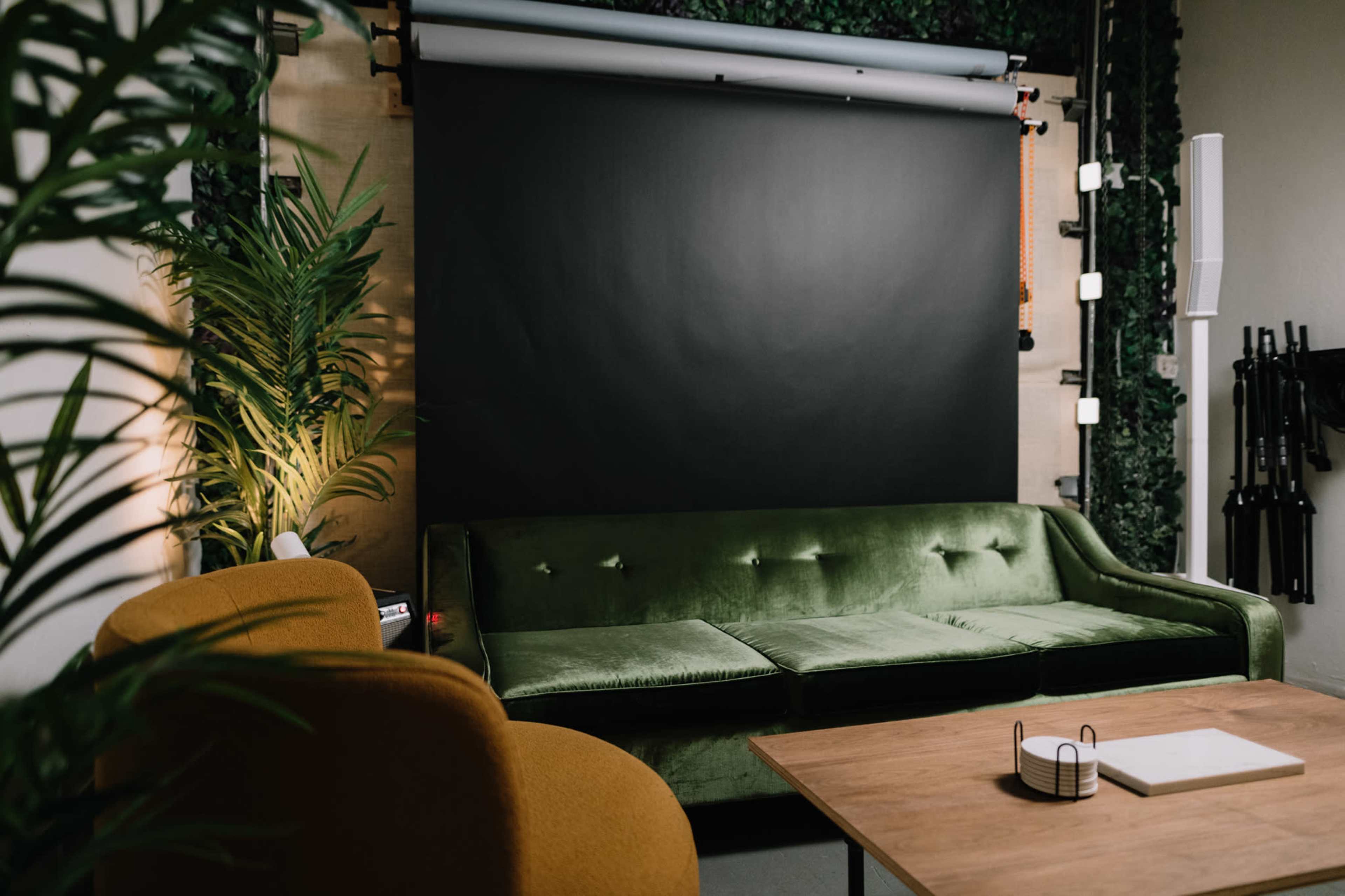 The image shows a cozy studio space featuring a green velvet sofa, an orange armchair, a large black backdrop, and plants on the walls.
