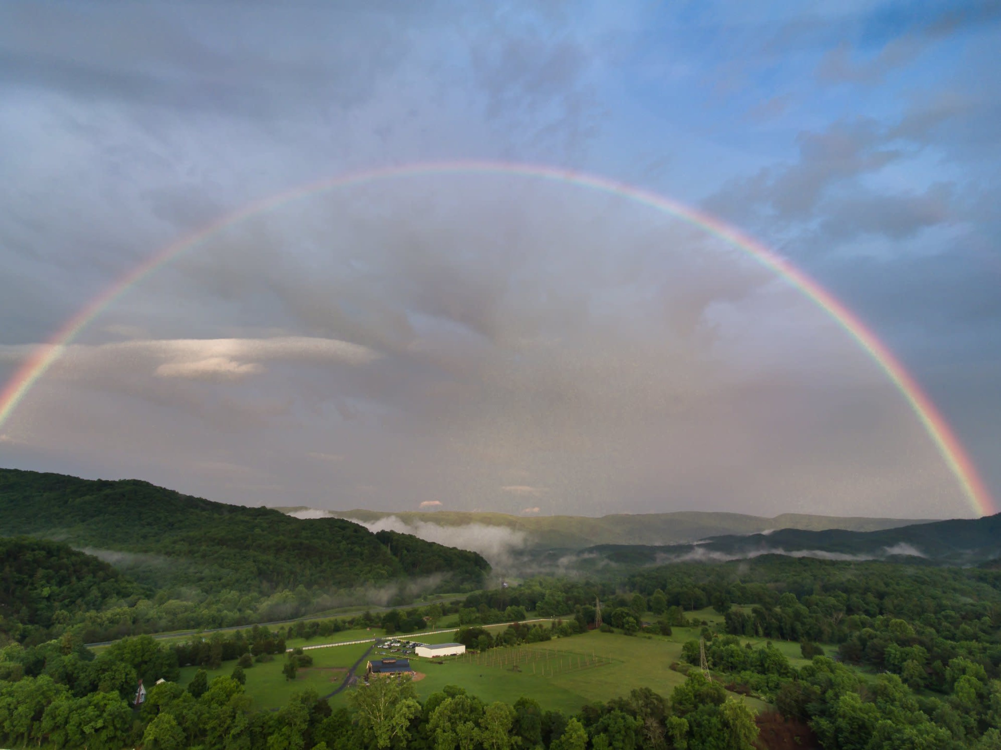 A wide rainbow arcs over a lush green landscape with rolling hills and a white building in the foreground.