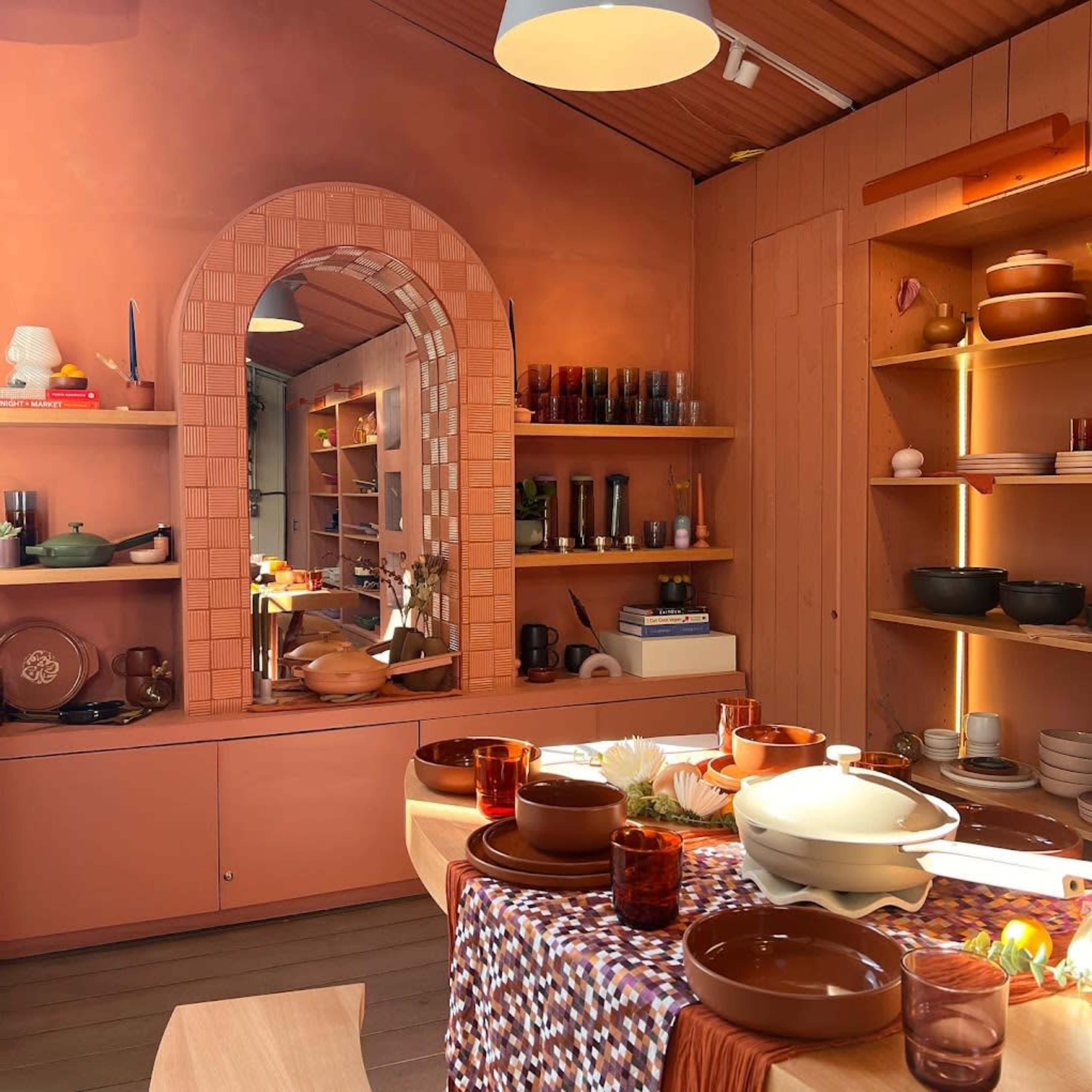 The image shows a warm-toned kitchen featuring an arch-shaped mirror, shelves with various dishes and glassware, and a table set with a colorful tablecloth and dinnerware.