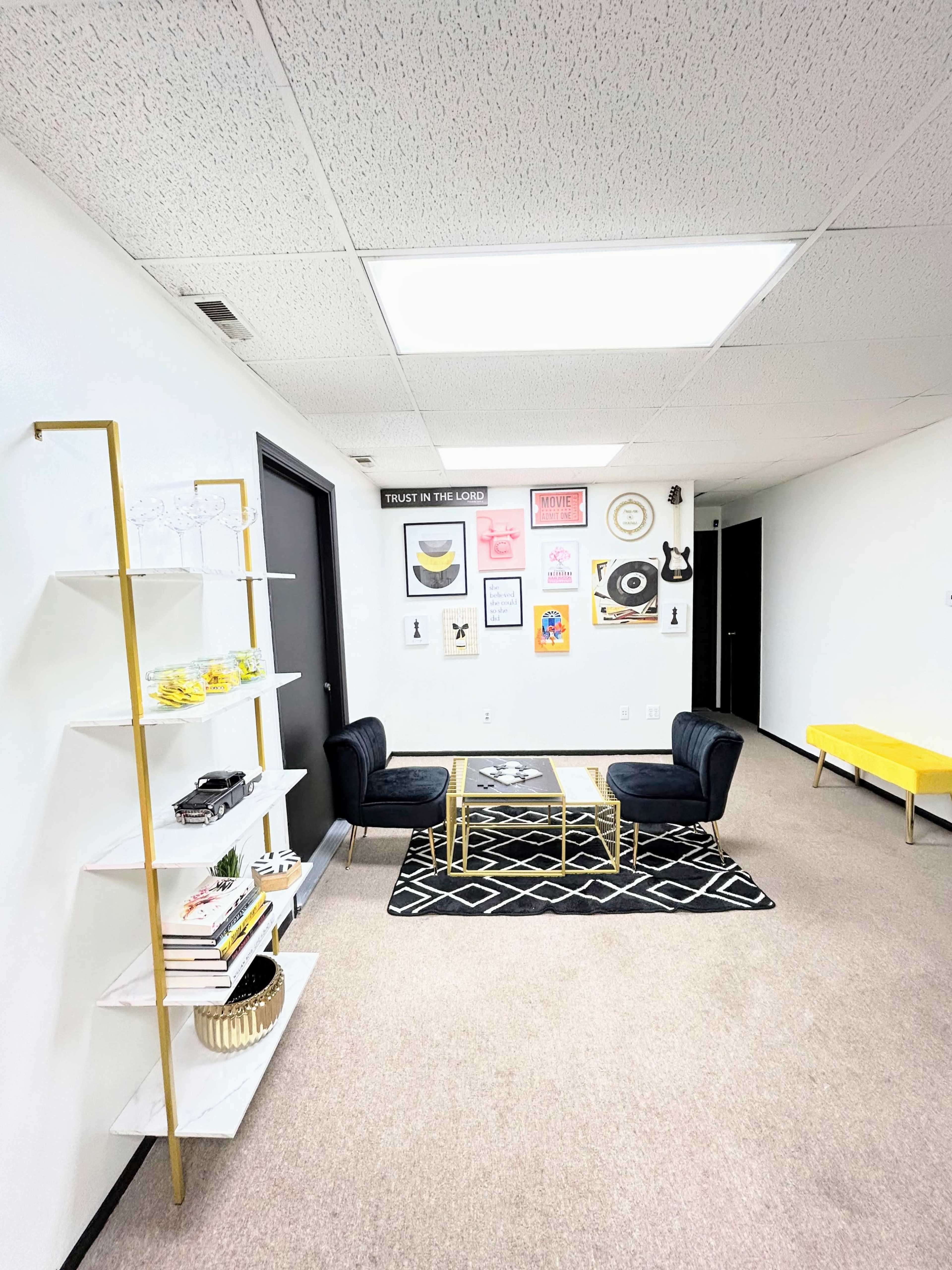 A modern office space with a black carpet, a coffee table surrounded by two black chairs, and a wall adorned with various framed art pieces.