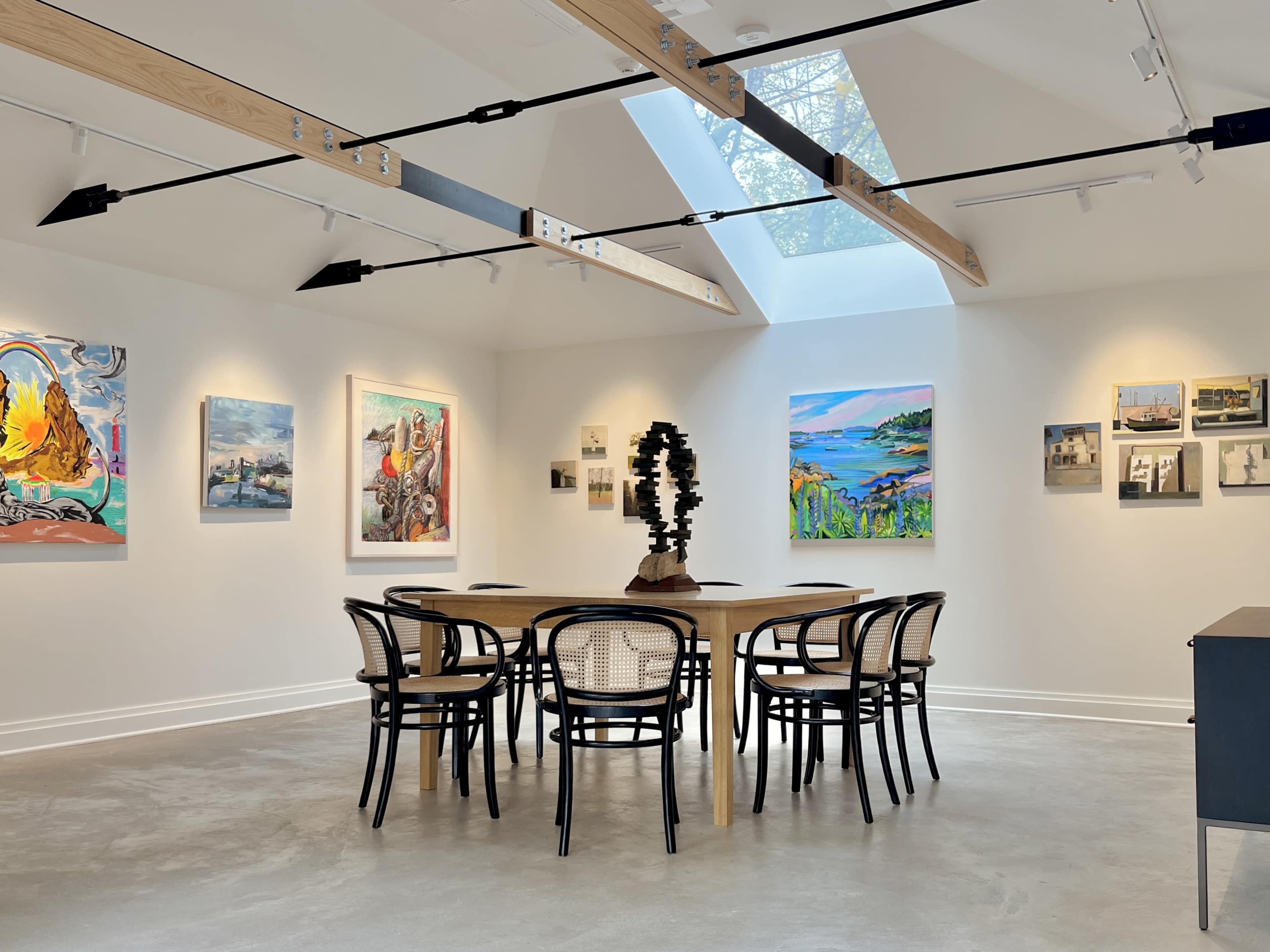 The image shows a modern art gallery featuring a circular wooden table surrounded by eight black chairs, with various pieces of colorful artwork displayed on the walls.