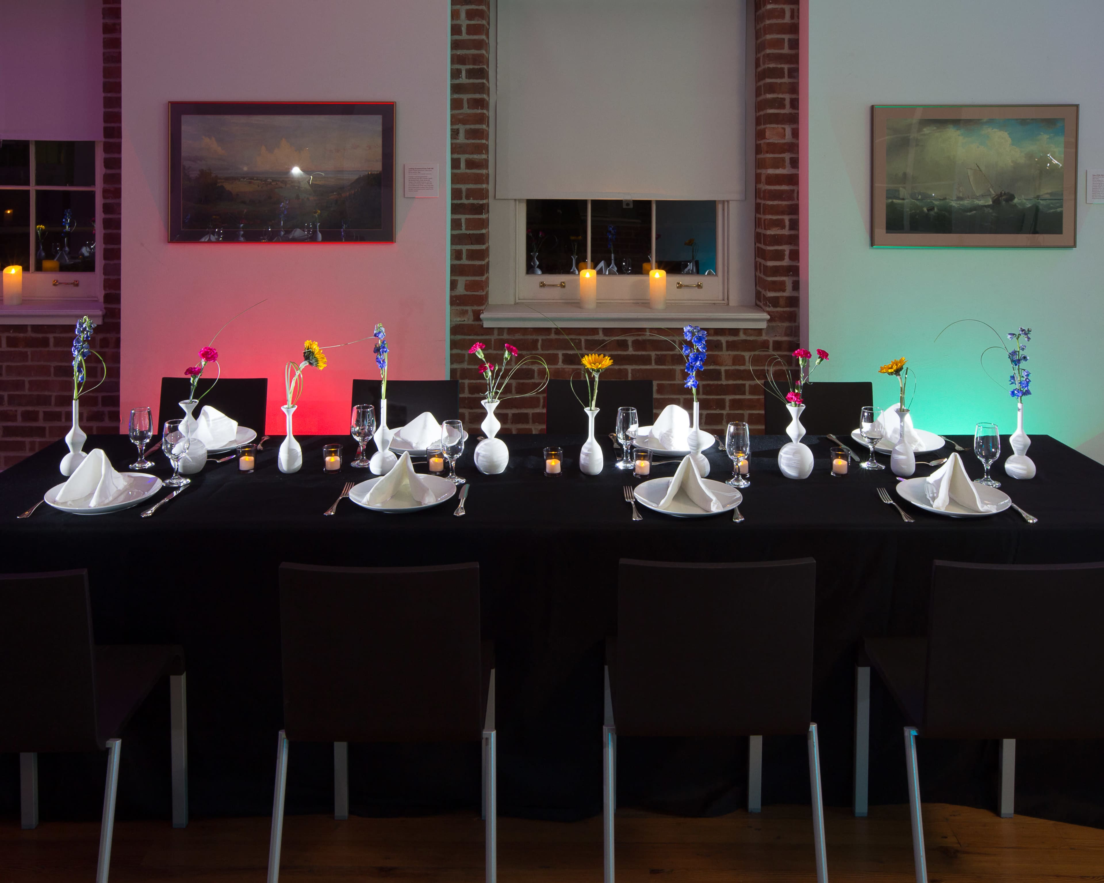 A long dining table is set with white tableware and decorated with small vases holding flowers, surrounded by black chairs in a well-lit room.