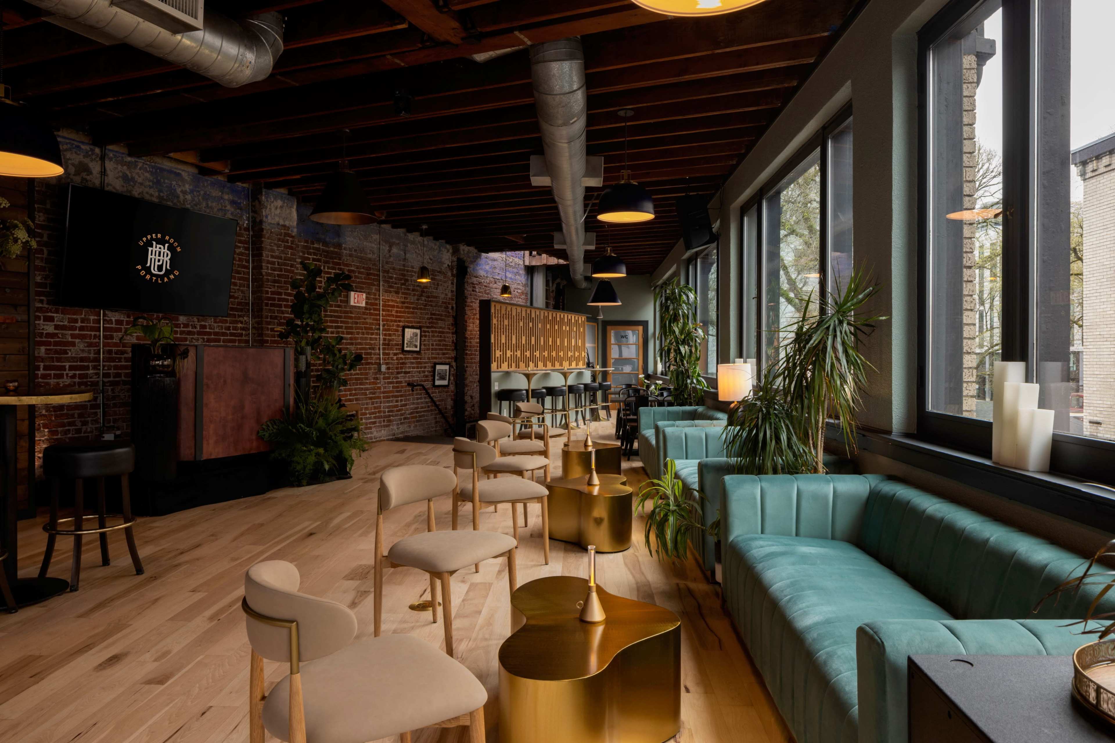 The scene shows a modern lounge with a mix of seating options, large windows, exposed brick walls, and wooden beams.