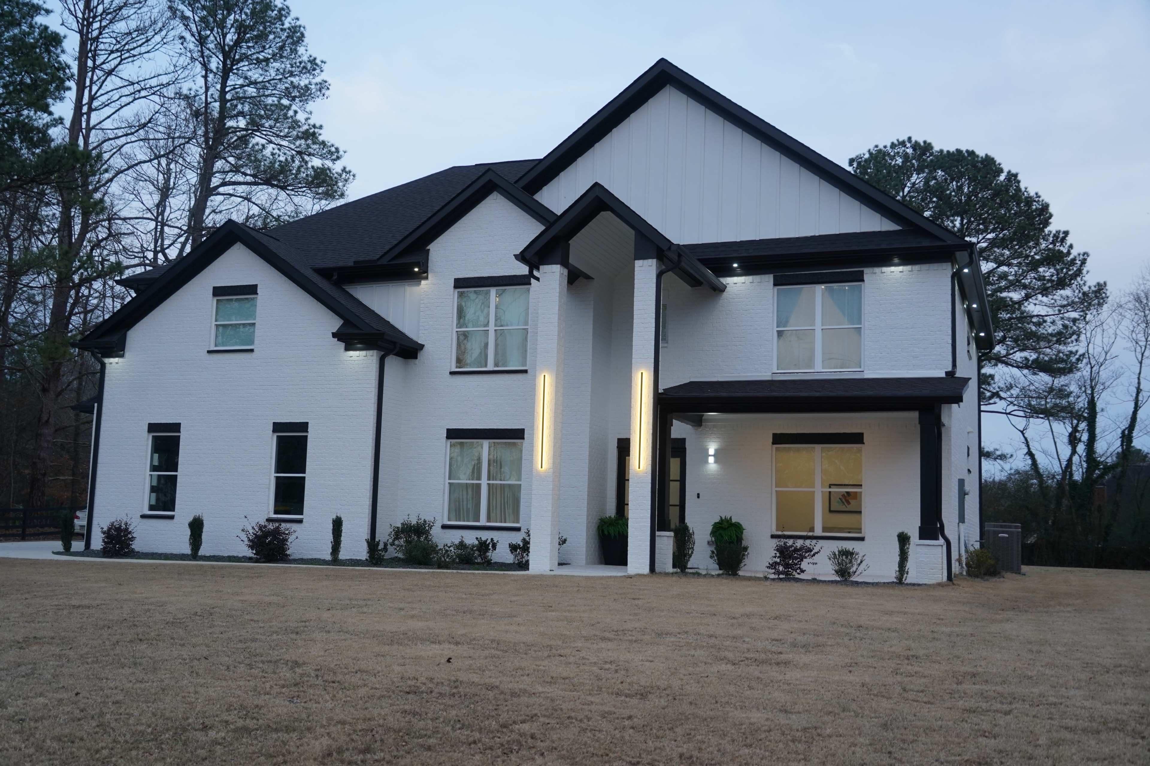 A modern two-story white house with large windows and a symmetrical design is situated on a grassy lawn surrounded by trees.
