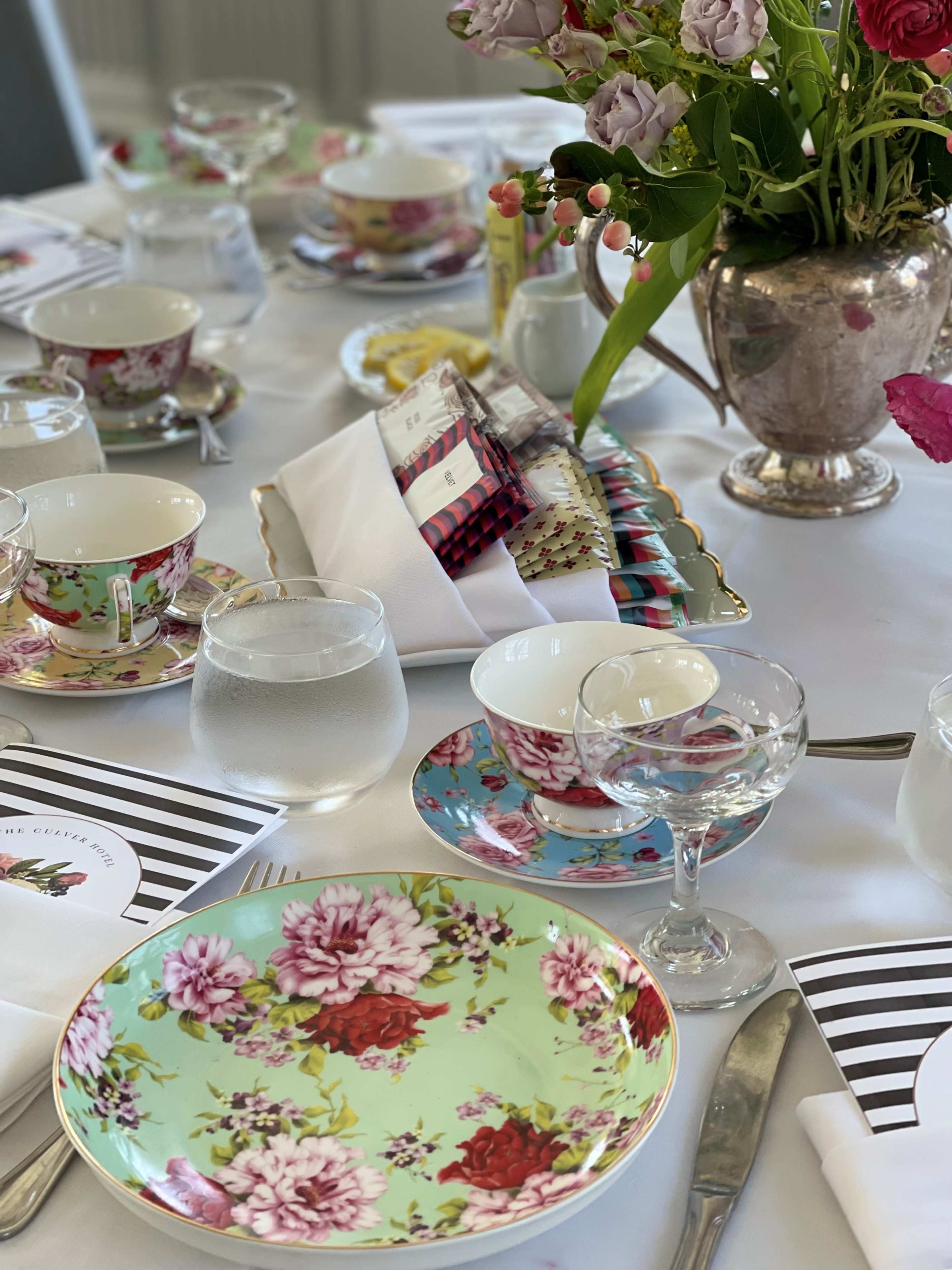 A beautifully arranged dining table features floral-patterned china, glassware, and a silver vase filled with fresh flowers.