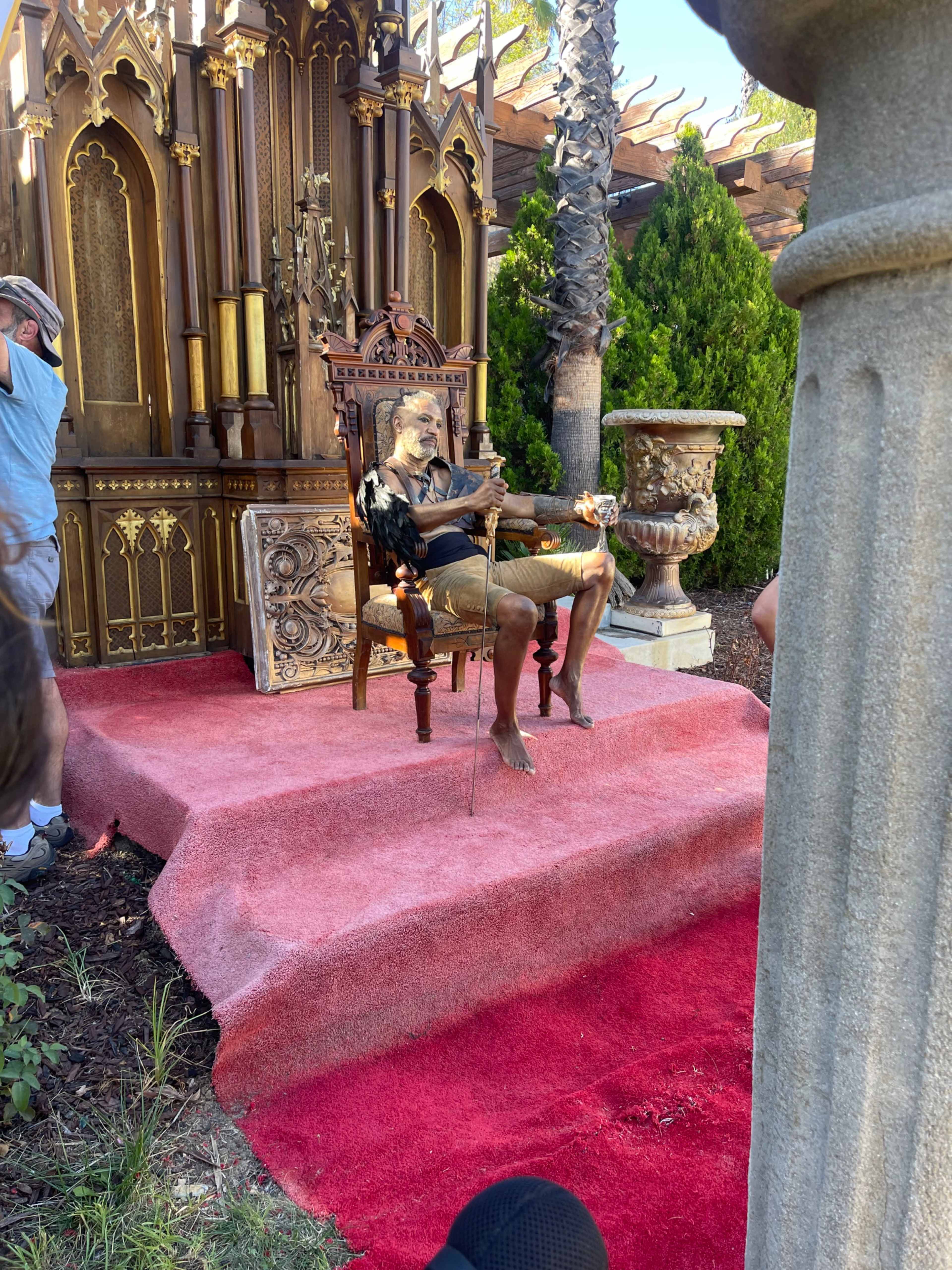 A statue of a seated figure in casual attire is positioned on a decorative throne surrounded by lush greenery and ornate structures.