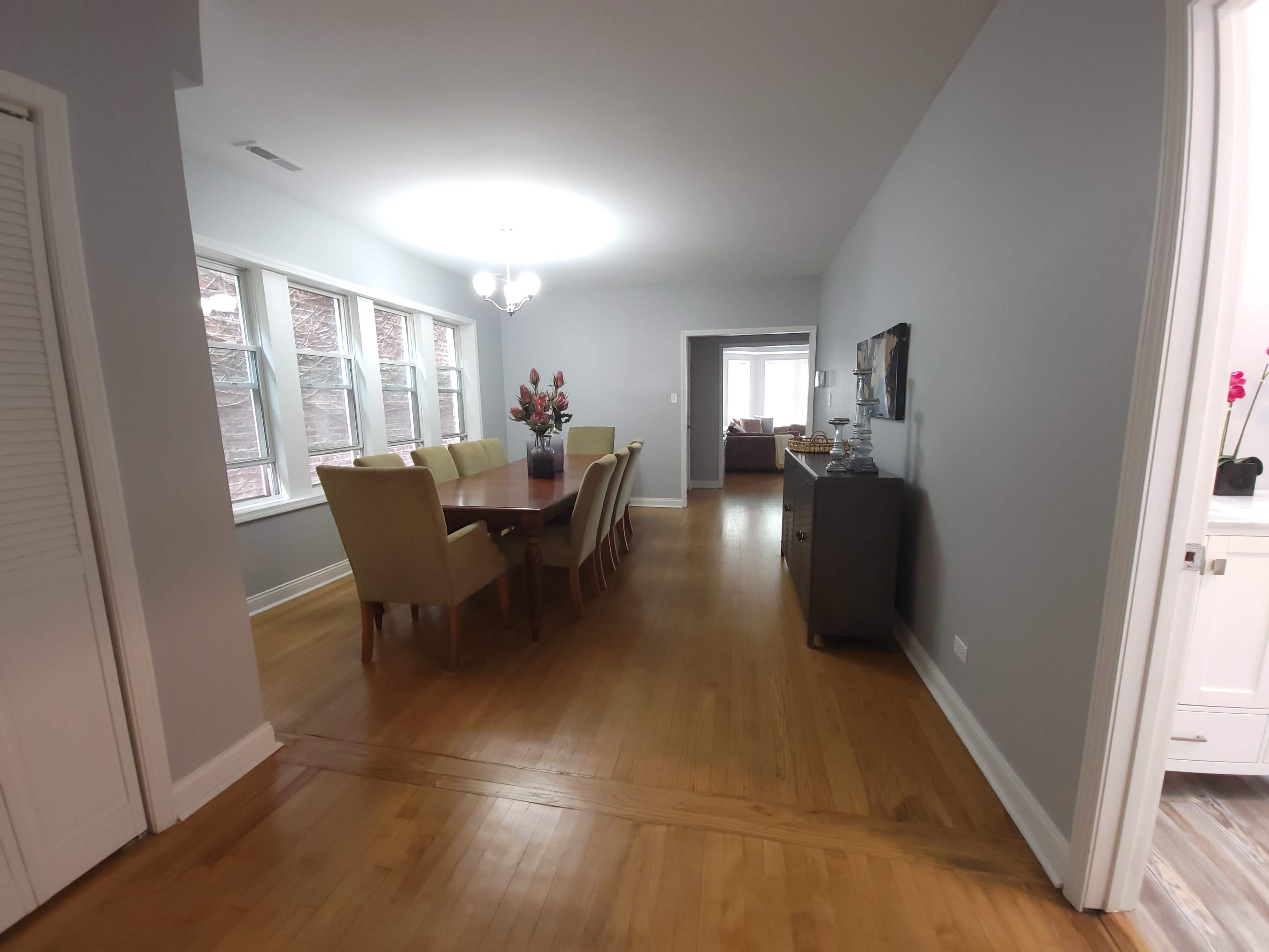 The image shows a spacious dining room with a long table surrounded by chairs, illuminated by a circular ceiling light, with a view into a living area beyond.