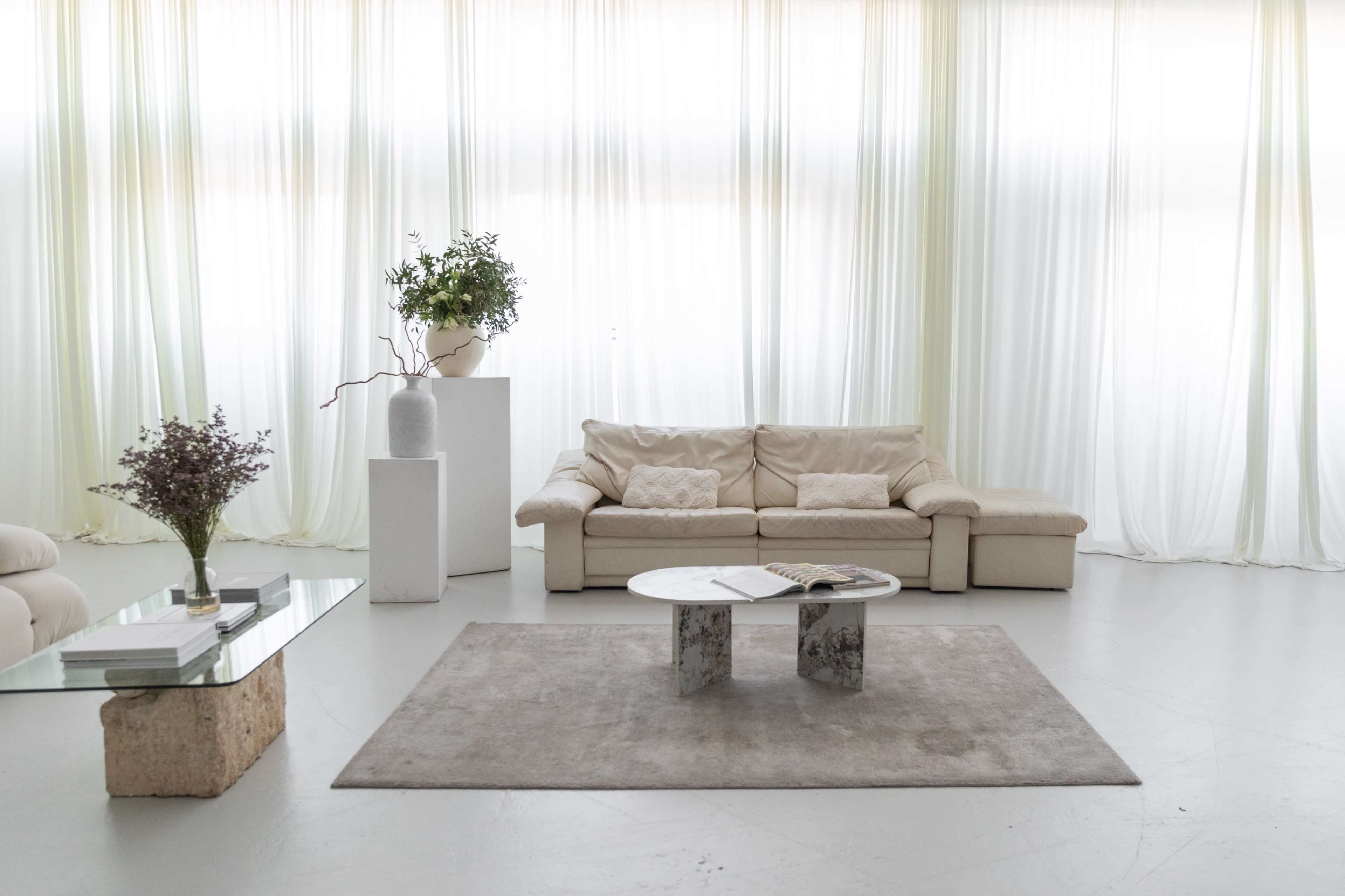 A modern living room features a beige sofa, a glass coffee table, and a light gray rug under large sheer curtains.