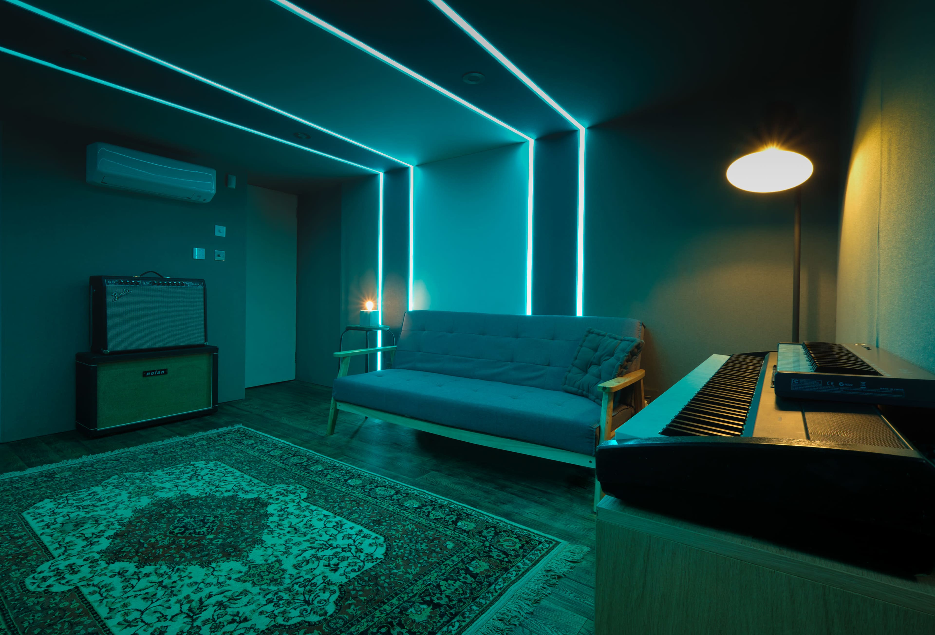 A dimly lit music studio features a blue-lit sofa, an electric keyboard on a wooden table, an amplifier in the corner, and a patterned rug on the floor.