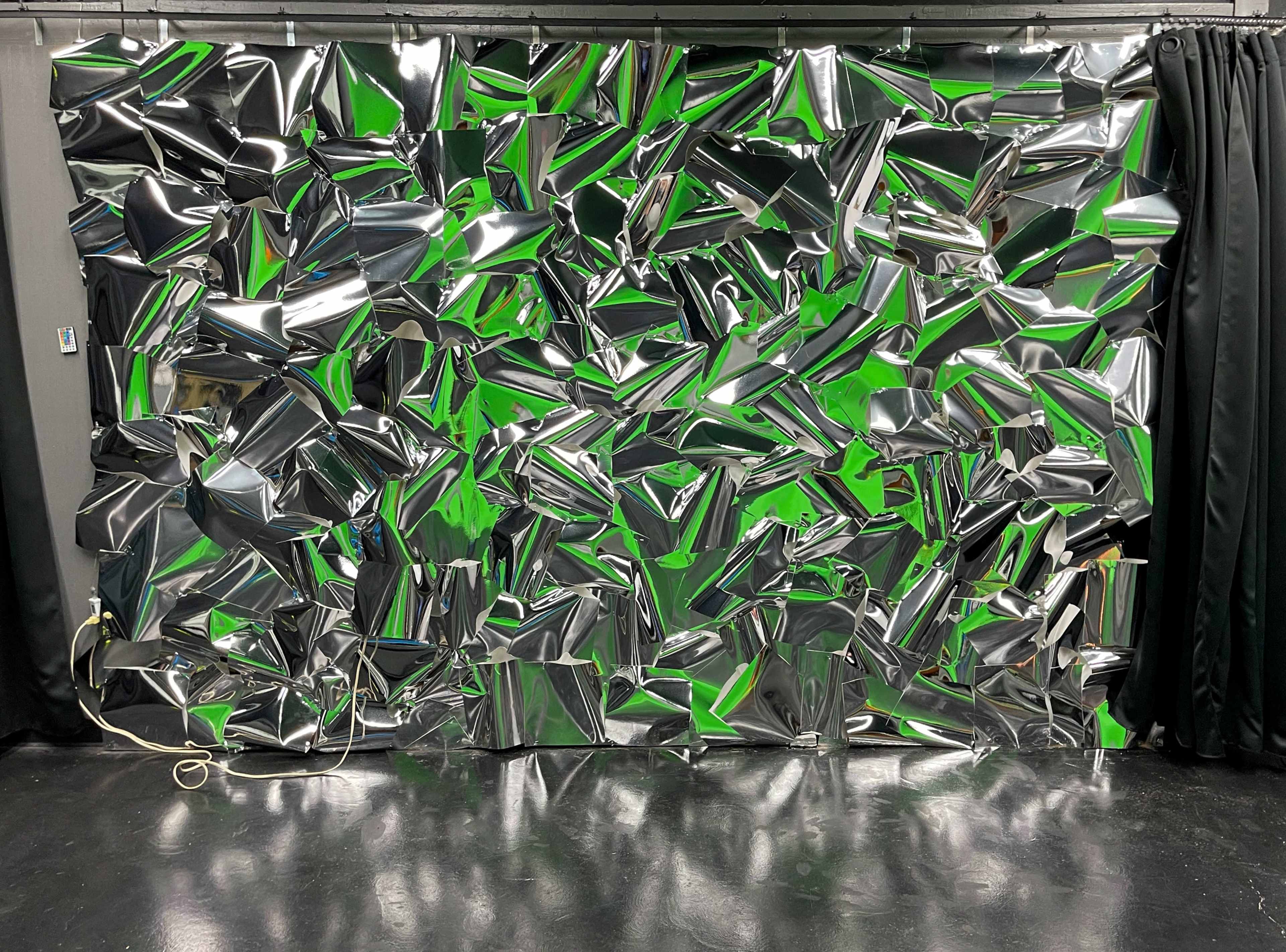The image features a wall covered with crumpled metallic foil in various shades of green and silver, with black curtains on either side.